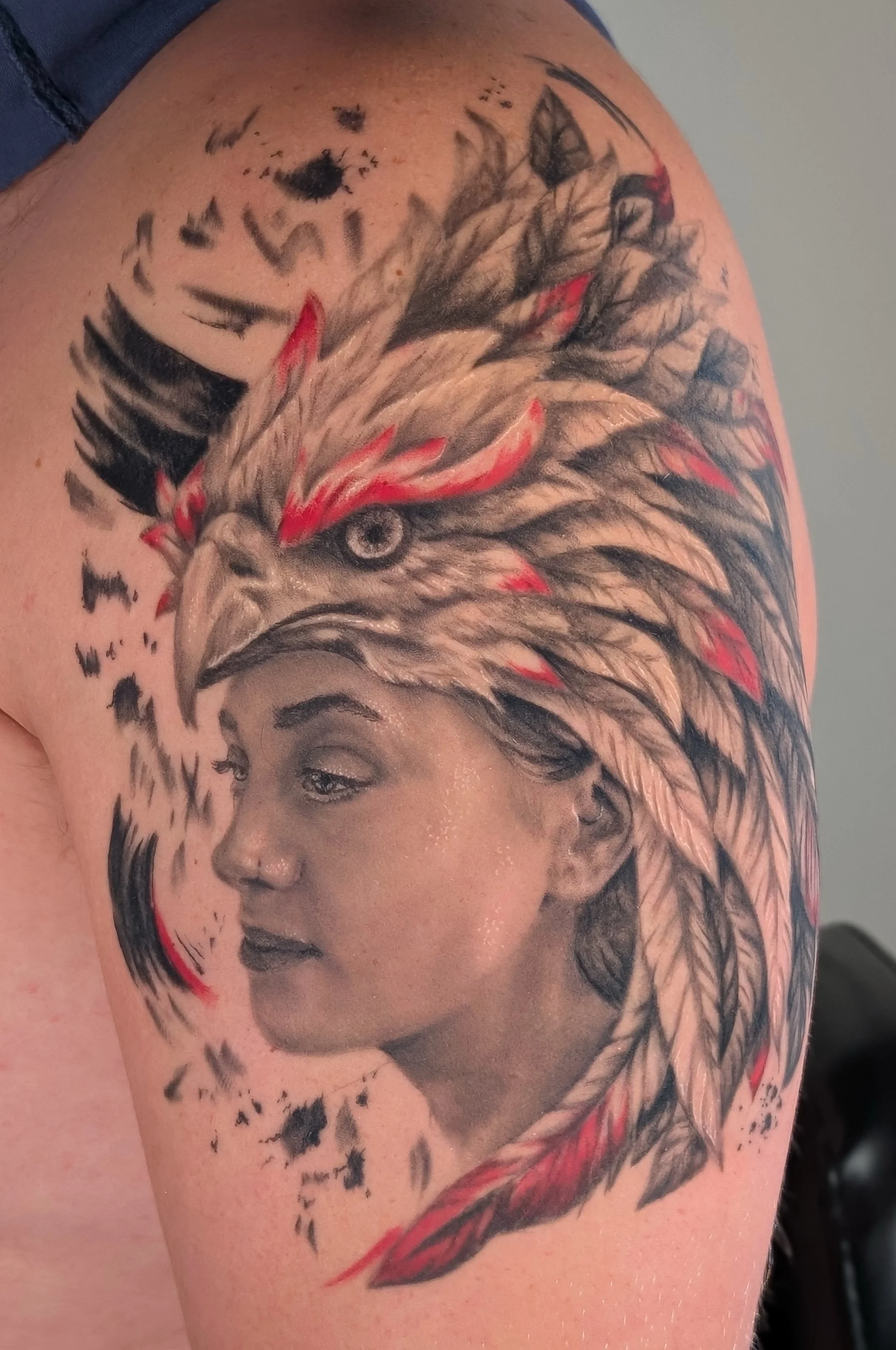 Tattoo of a woman's face with a detailed depiction, including shading for realism, and an eagle's head with red accents and feathers above her, on a person's upper arm.