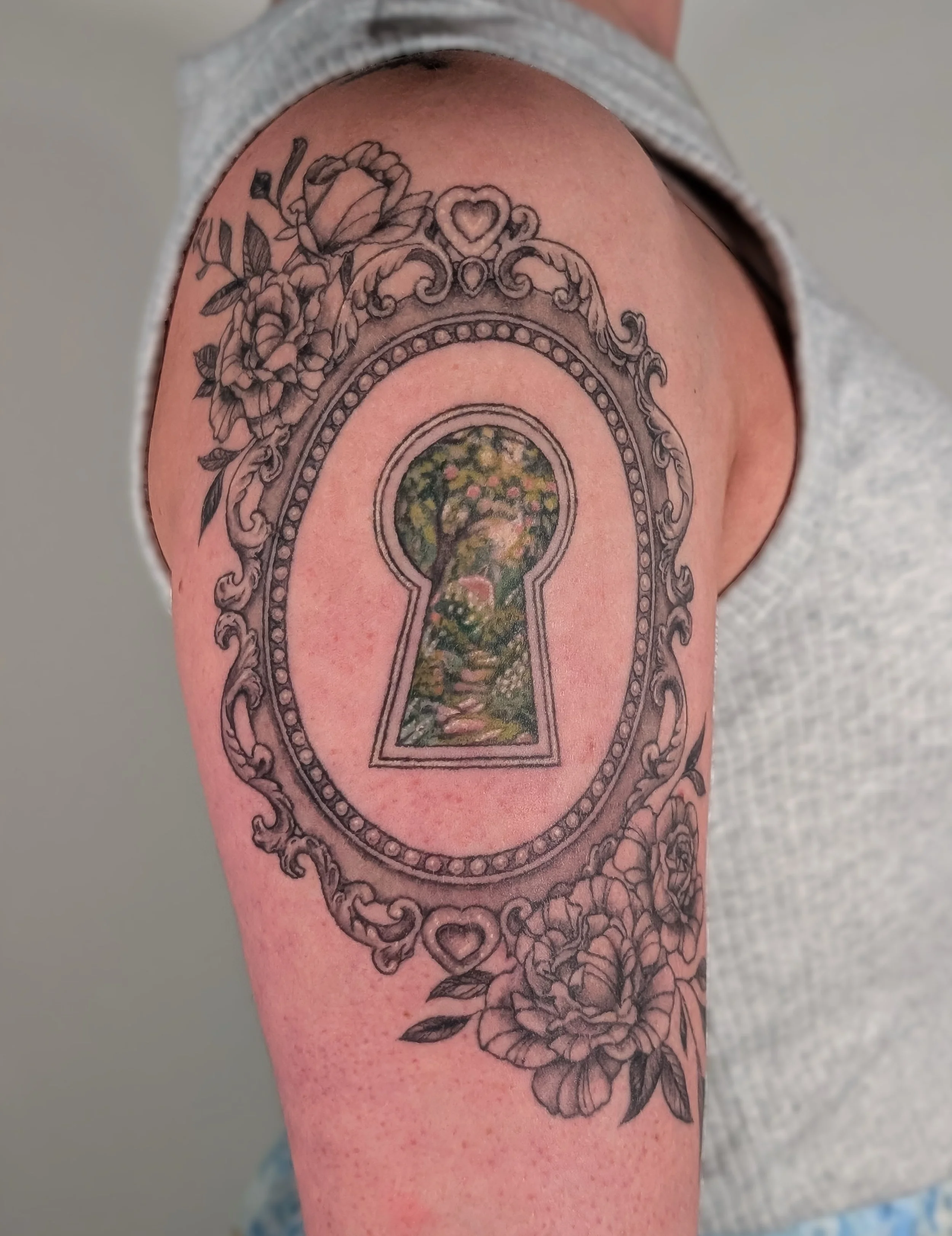 Tattoo of a decorative mirror frame with roses and a heart at the top, surrounding a keyhole-shaped scene of a tree with a path and landscape inside.