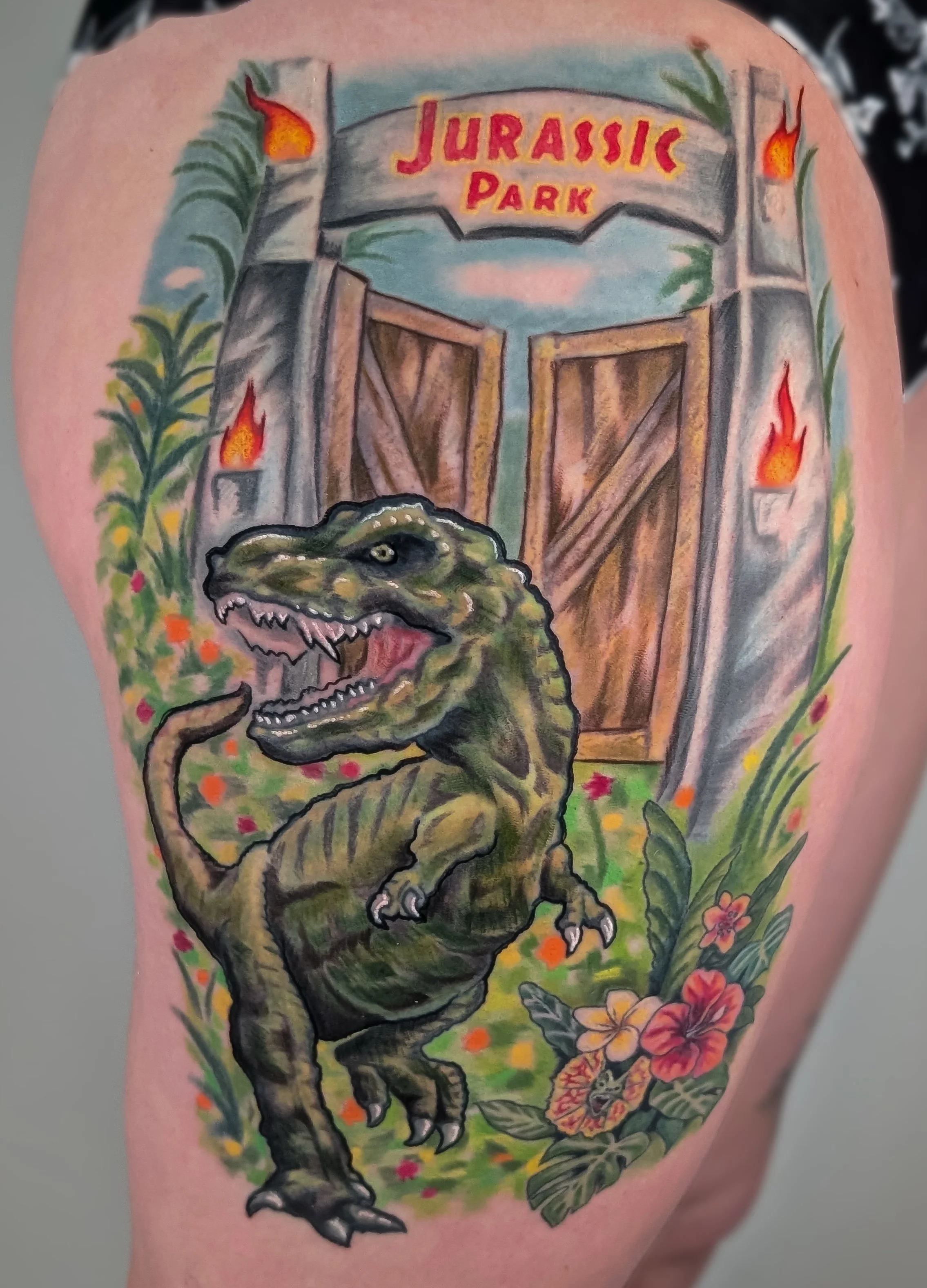 Colorful tattoo of a dinosaur in front of a Jurassic Park gate with flowers and flames.