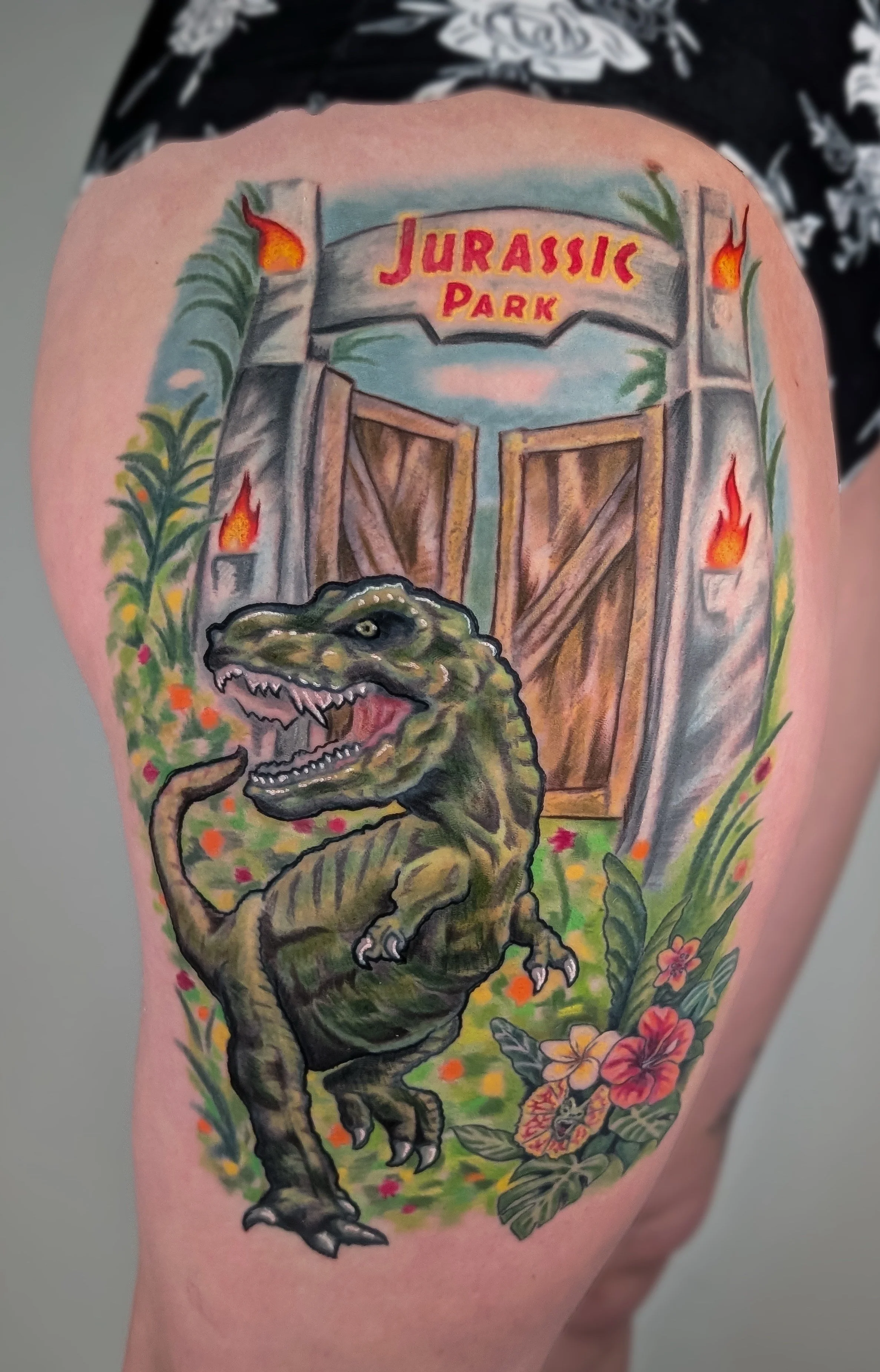Reworking and coverup of an existing dinosaur tattoo and gate.  Caitie added the greenery and flowers, added depth to the dinosaur, and added the Jurassic Park logo to honor the client's love for the movie.  The tattoo is colorful and takes up most o