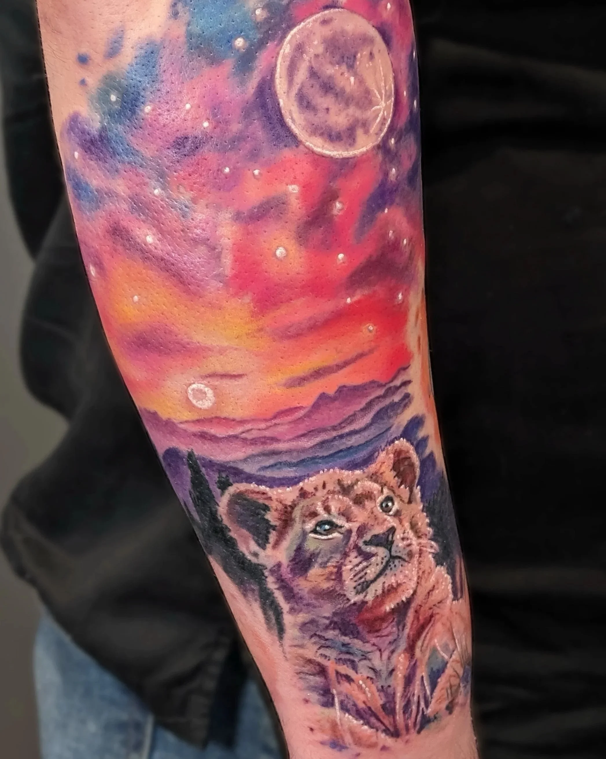 Colorful tattoo of a lion cub in a natural landscape with a sunset, mountains, and a starry sky with the moon.