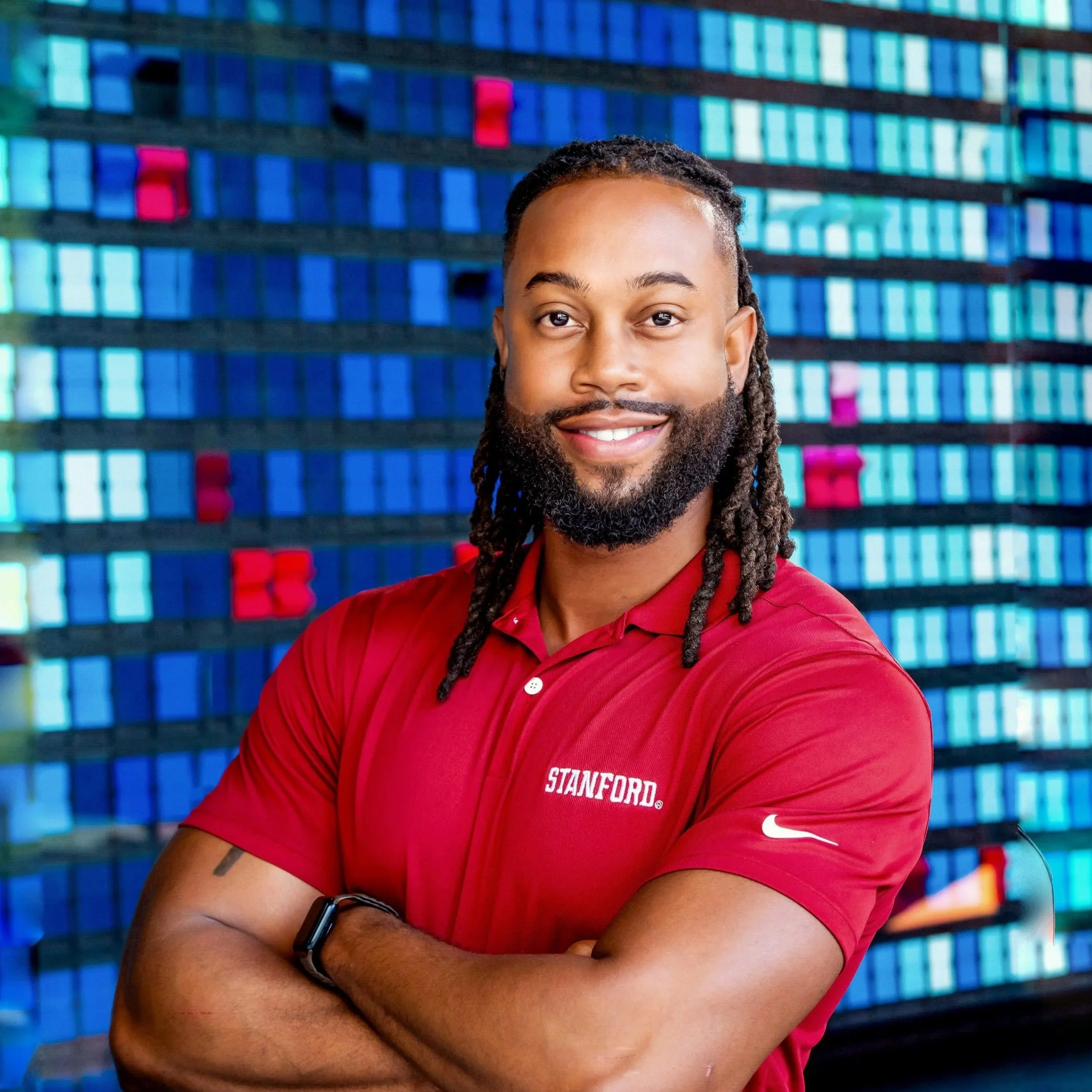Smiling person with a beard and dreadlocks in a red shirt, standing in front of a colorful patterned background.