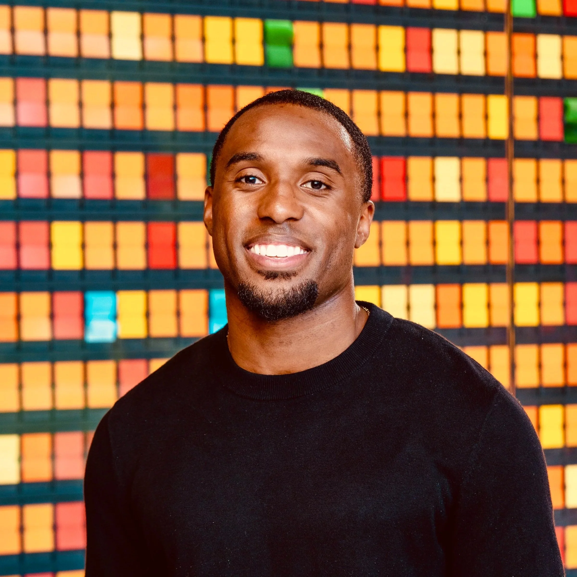 Smiling person with short curly hair wearing a black sweater against a plain background.