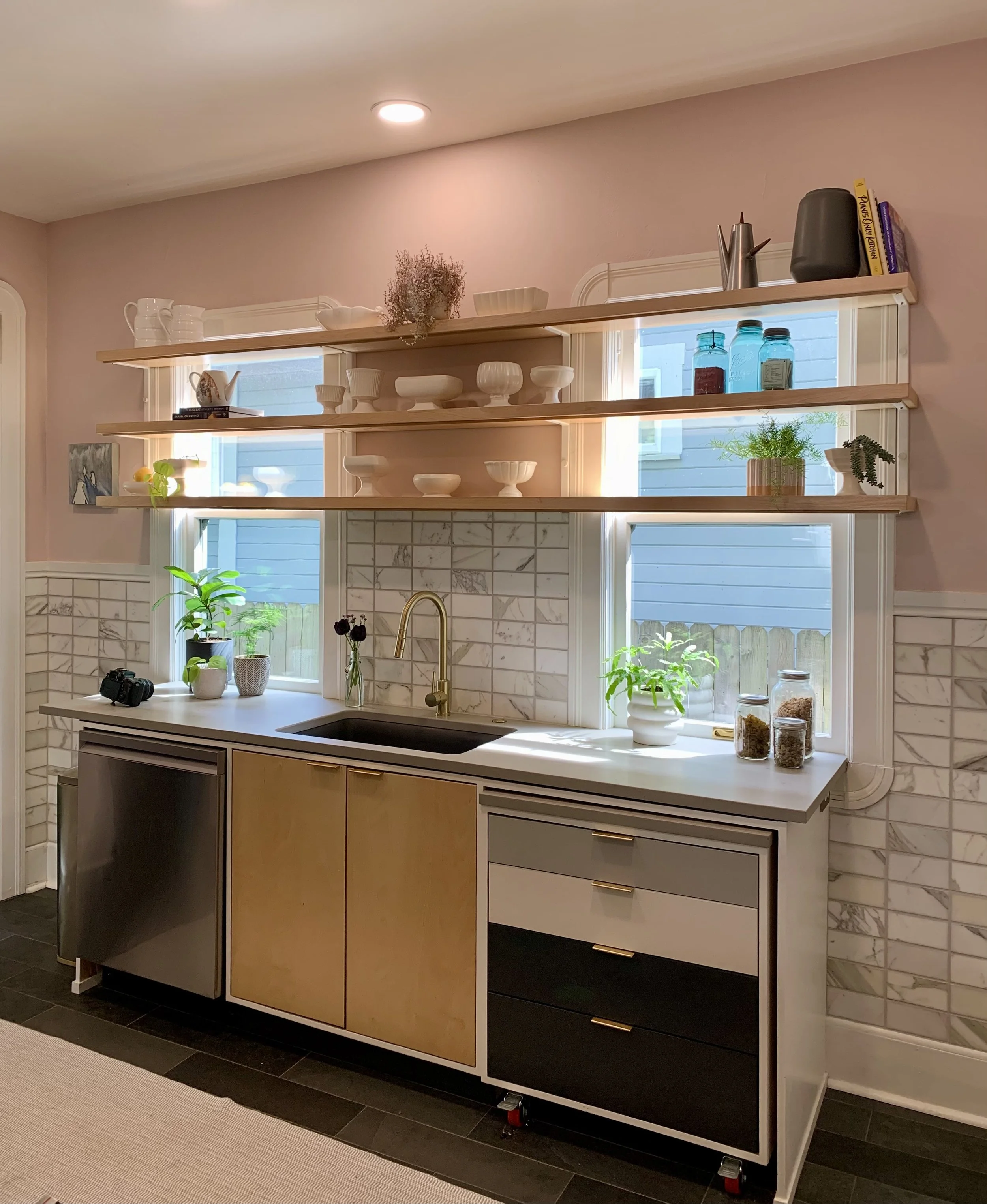 Modern shelving, countertops, and drawers in a modern Portland kitchen. Kitchen area with wooden open shelves, plants, and jars above a white countertop with a sink, gold faucet, and beige cabinetry, set against a pink wall and tiled backsplash.