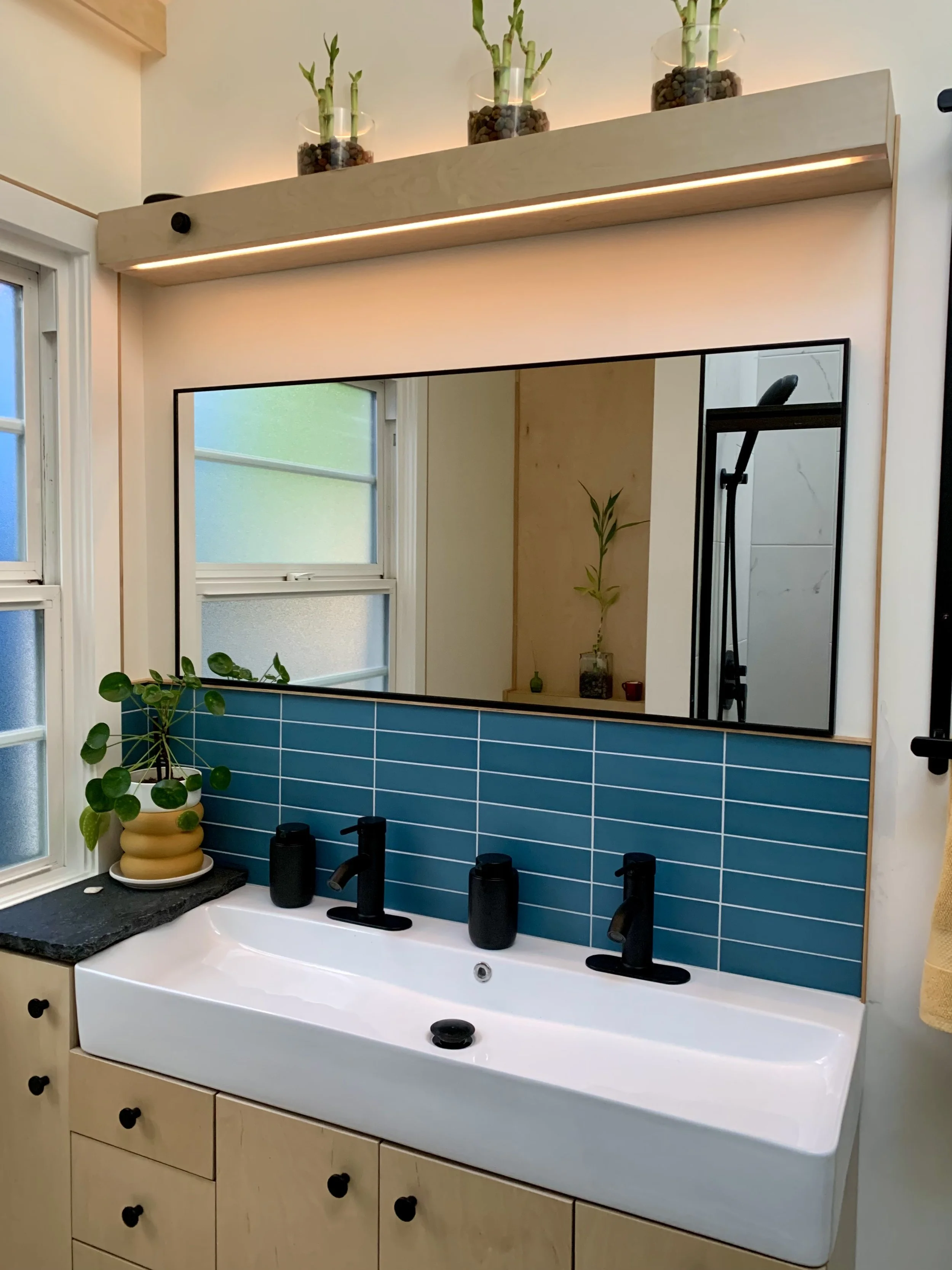 Custom-built bathroom vanity in Portland, OR. Stylish, modern bathroom sink with a large mirror above it, blue tiles as a backsplash, and a potted plant on the left.