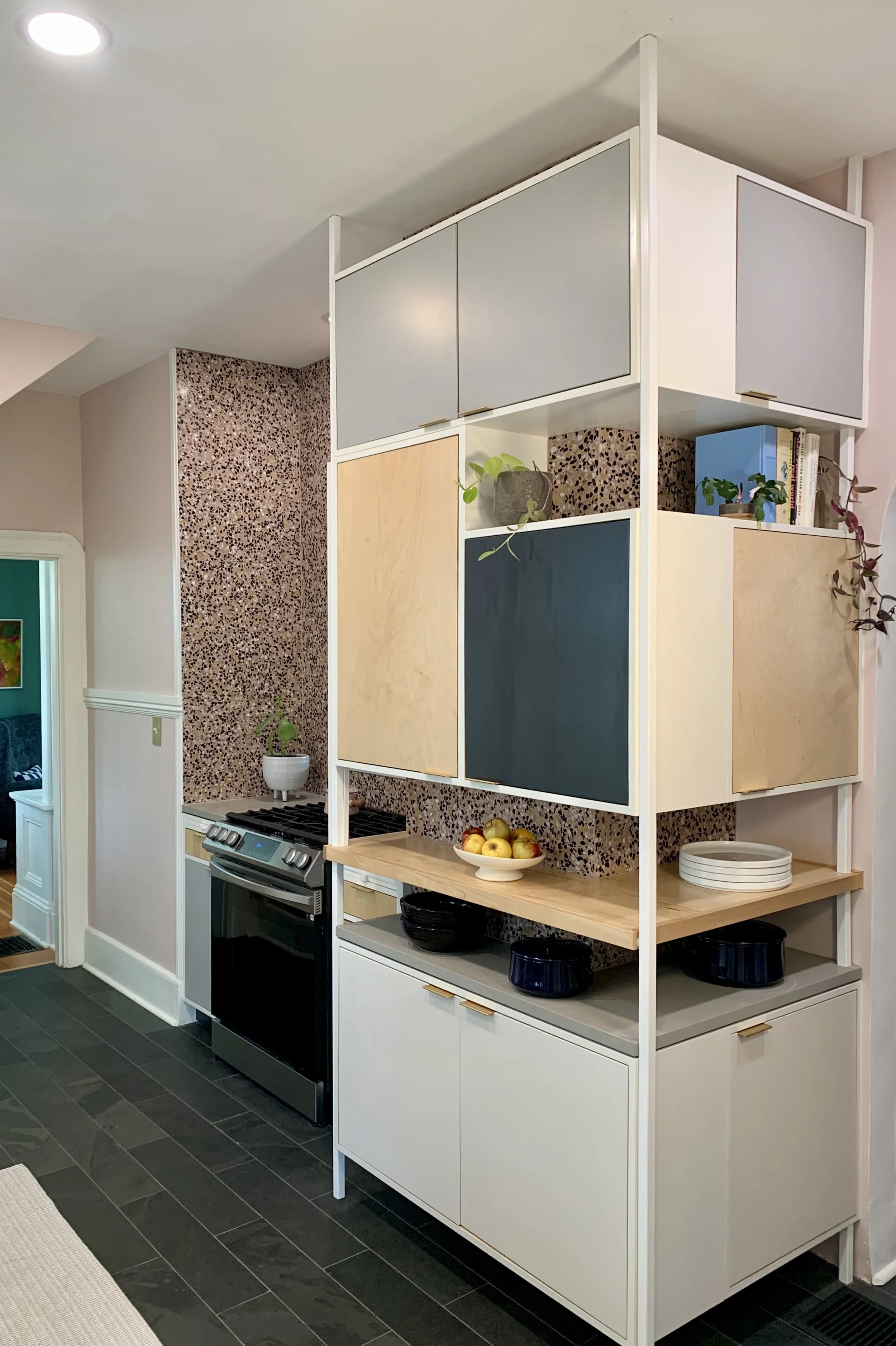 Custom cabinetry for additional kitchen storage space. Modern kitchen shelving unit with colorful cabinets, a countertop with bowls of fruit, and potted plants, adjacent to a stove, in a kitchen in Portland, Oregon.