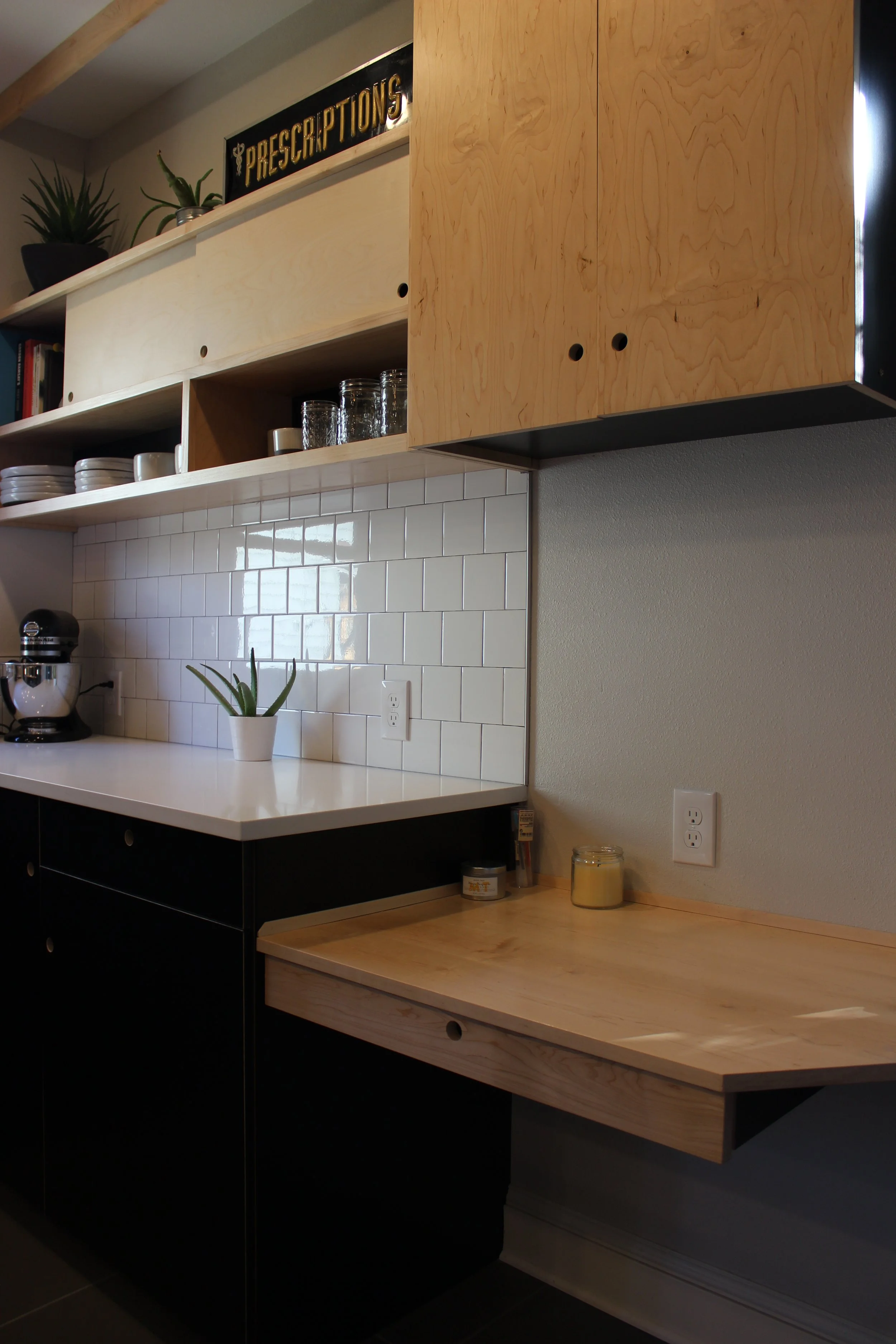 Custom kitchen cabinets and counter in Portland. Part of a modern kitchen with black and natural wood cabinets, white tiled backsplash, a potted succulent plant, glass jars, cups, and a small countertop with a candle and a jar.