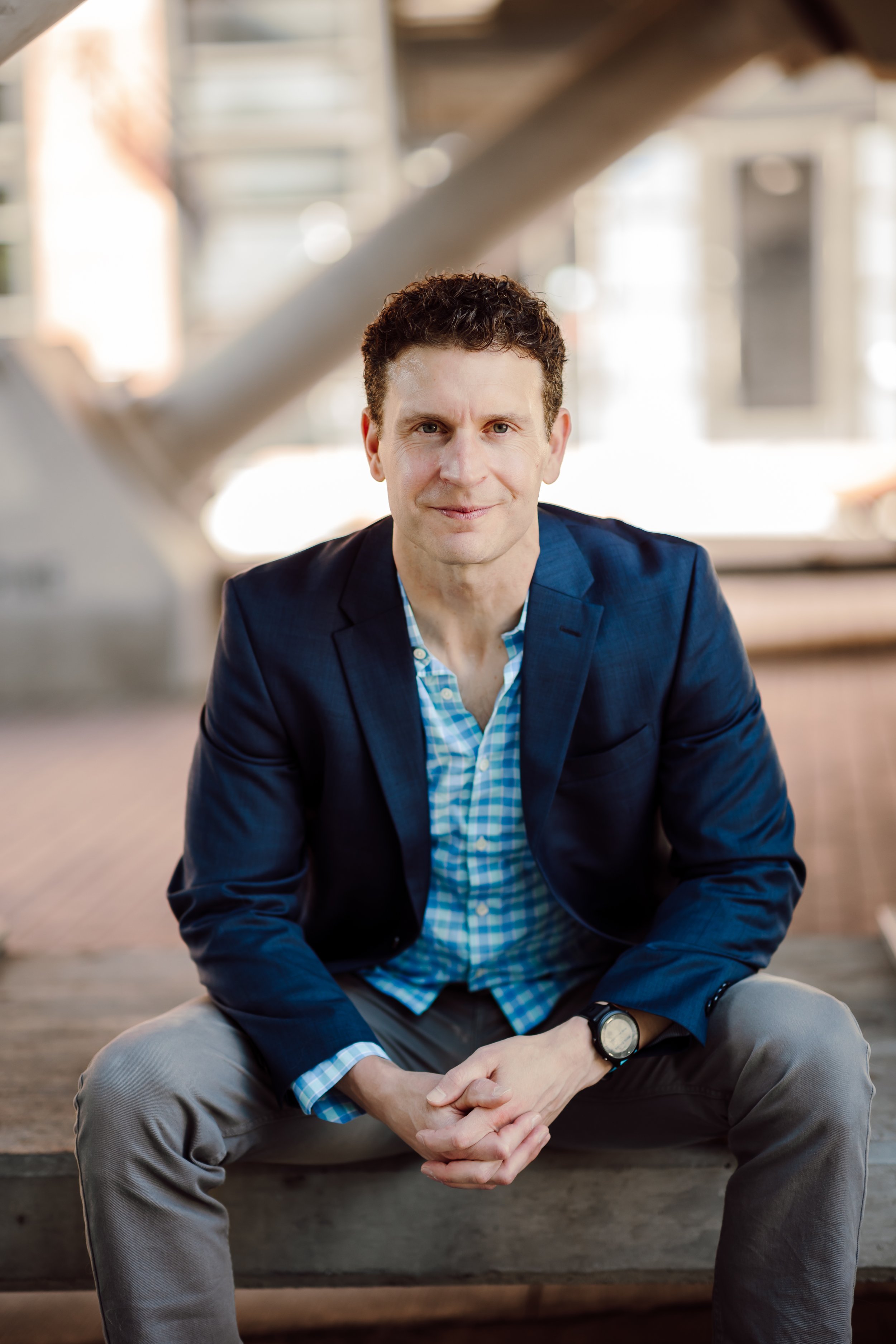 A man with curly dark hair, wearing a navy blazer, light blue checked shirt, and gray pants, sitting on wooden steps with his hands clasped and a watch on his wrist, smiling softly at the camera in a bright, industrial-style setting.
