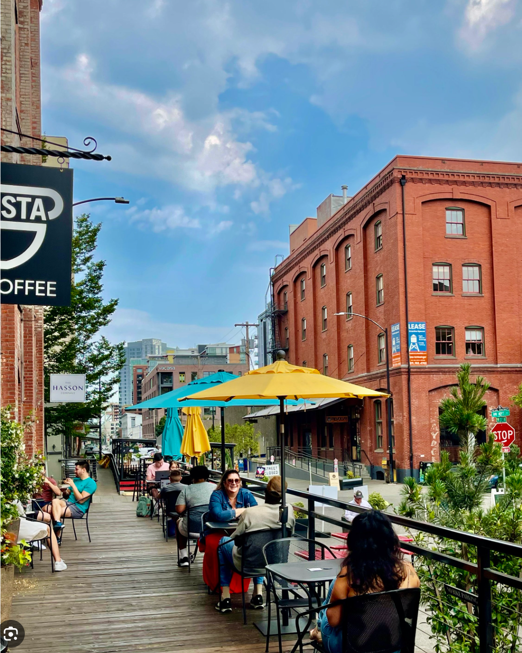 People sitting and socializing at outdoor tables on a wooden patio, with colorful umbrellas, next to a brick building in the Pearl District under a partly cloudy sky.
