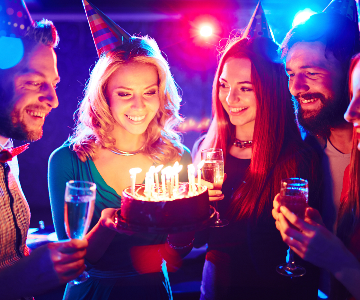 Group of friends celebrating birthday with cake, candles, party hats, and drinks at a party.