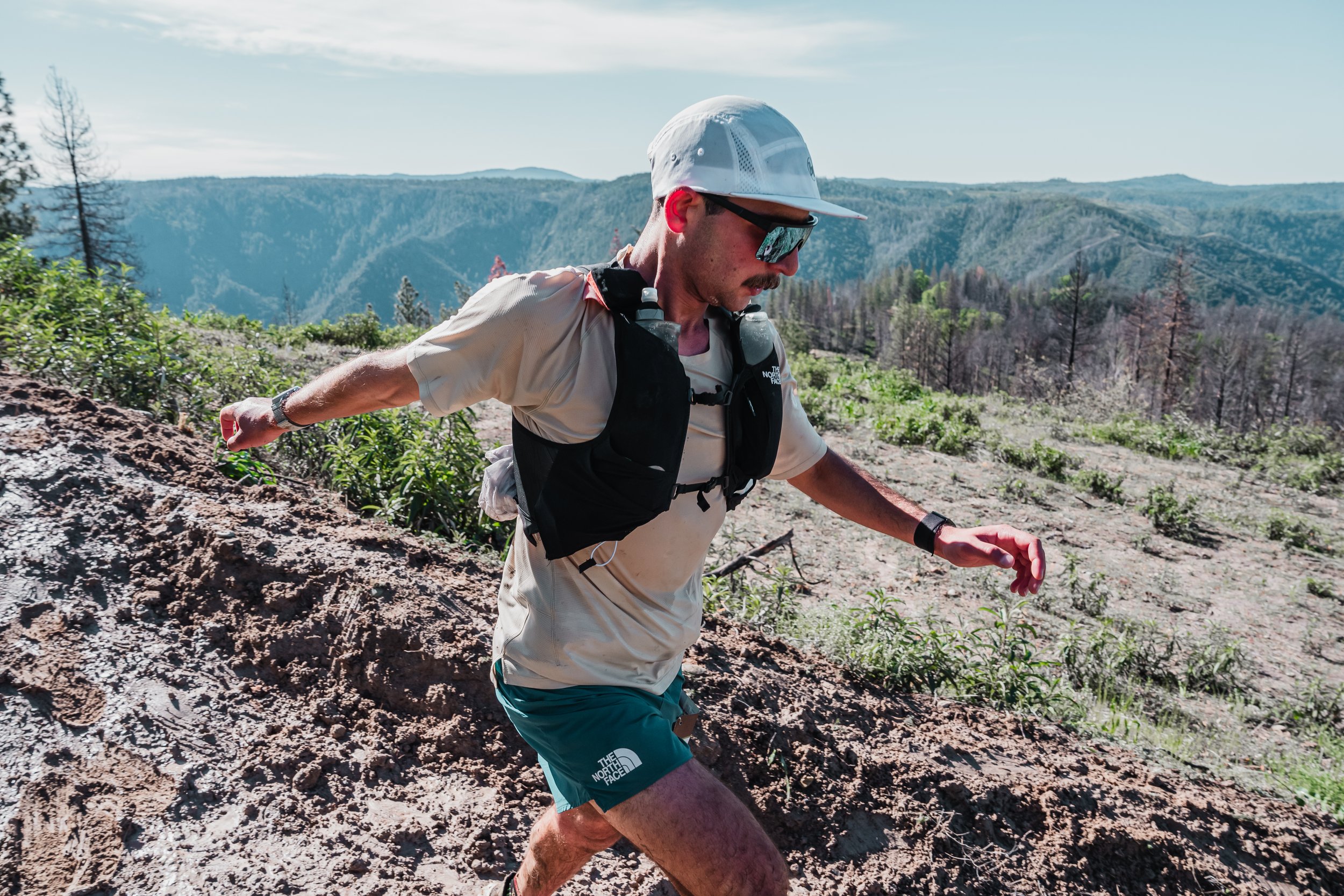 Trail runner wearing a hat and sunglasses, running through a mountainous landscape with greenery and dirt path.