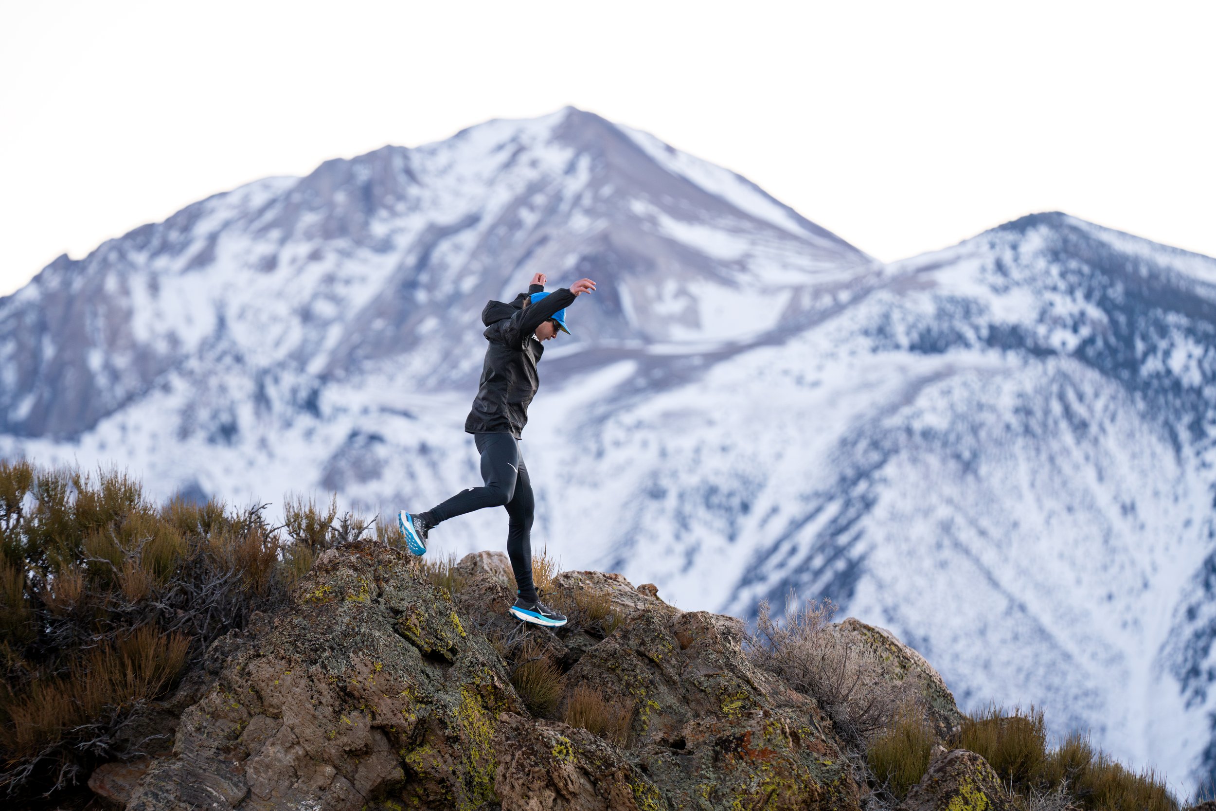 Person trail running on rocky terrain with snow-covered mountains in the background.
