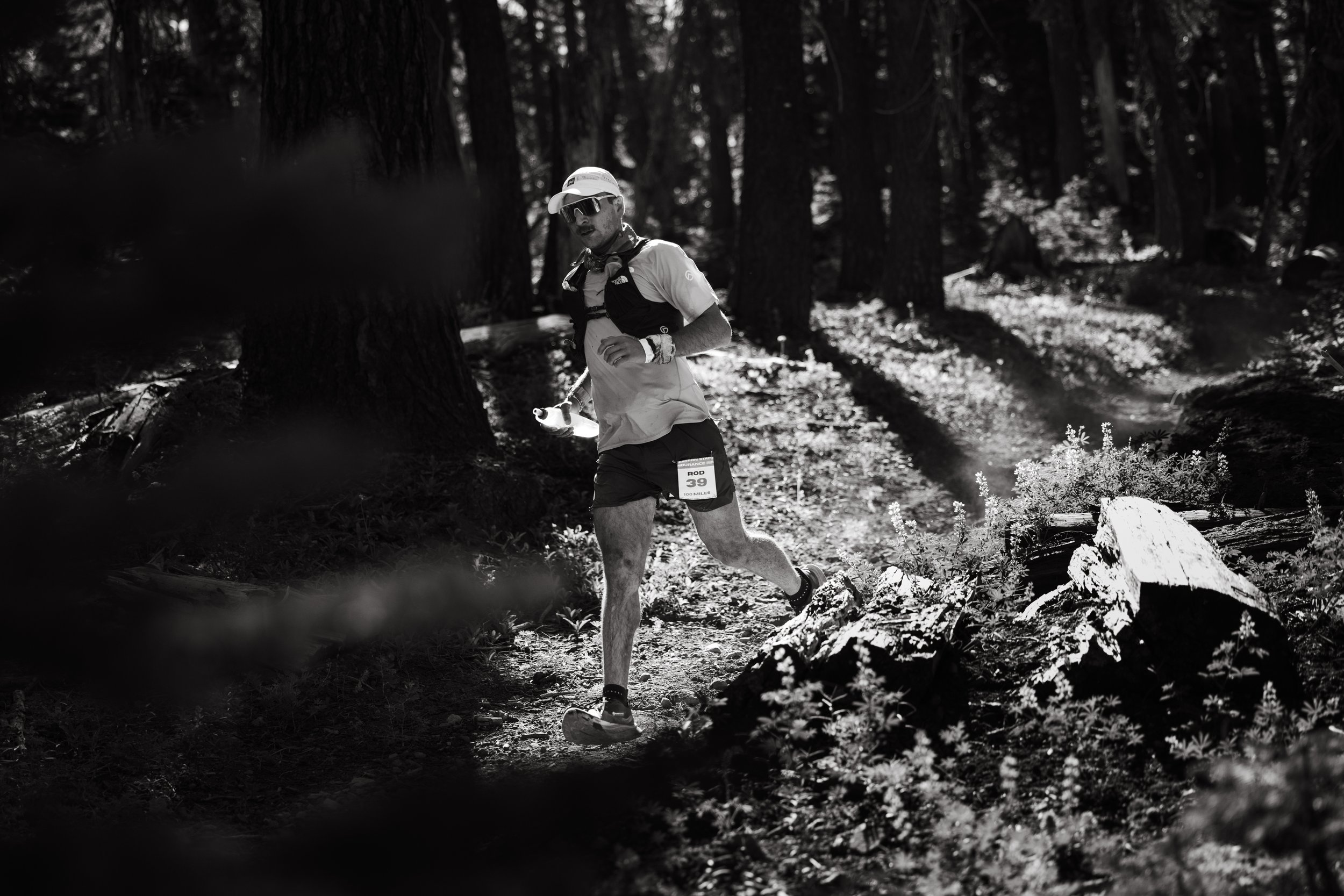 A monochrome image of a male trail runner wearing a race number 39, running through a forested trail. He is wearing a cap, sunglasses, and hydration gear. Sunlight filters through the trees, casting shadows on the path.