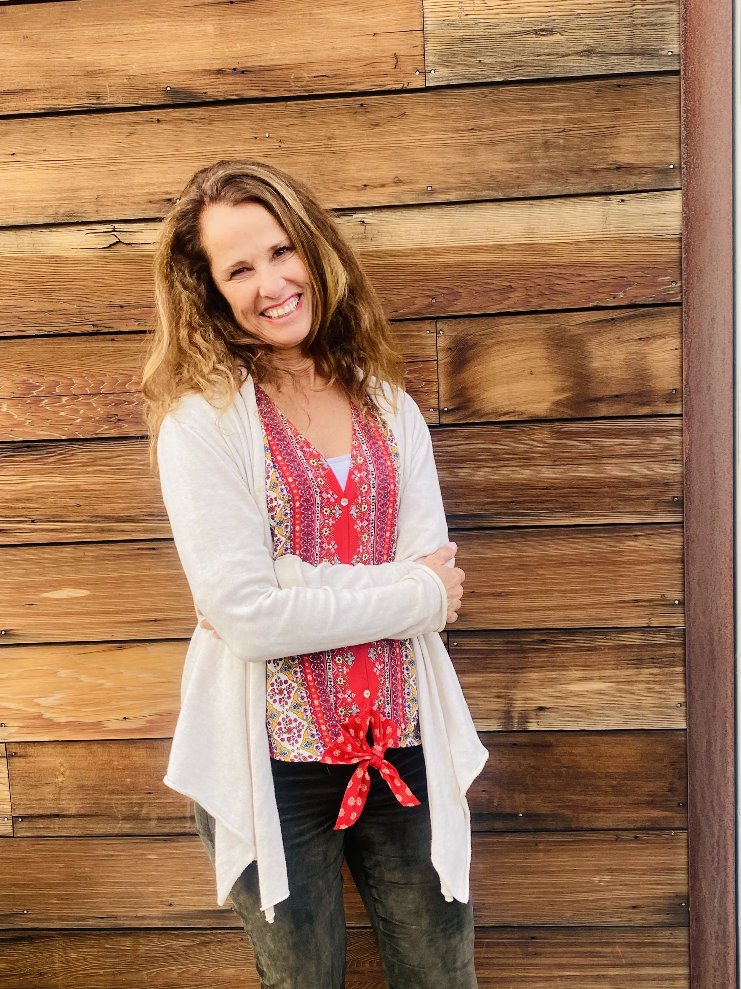 A woman with dark, curly hair smiling, wearing earrings and a necklace, indoors with a blurred background.