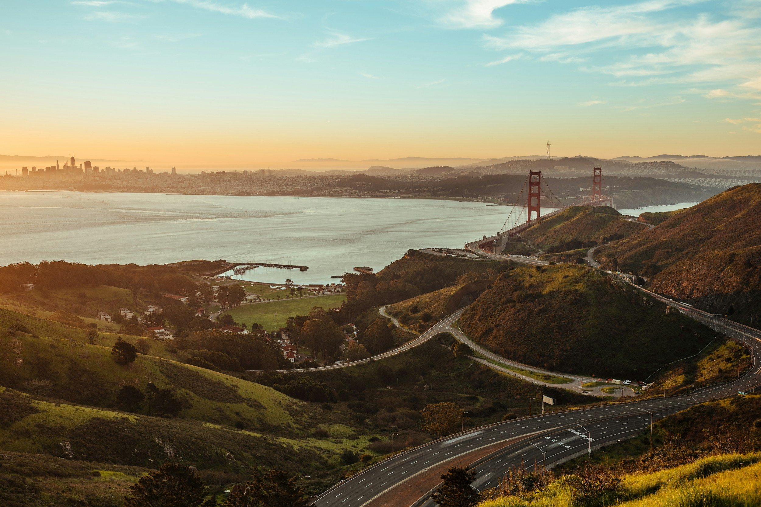 A scenic view of the Golden Gate Bridge in San Francisco during sunset, with winding roads on green hills in the foreground and the city skyline in the background across the bay.