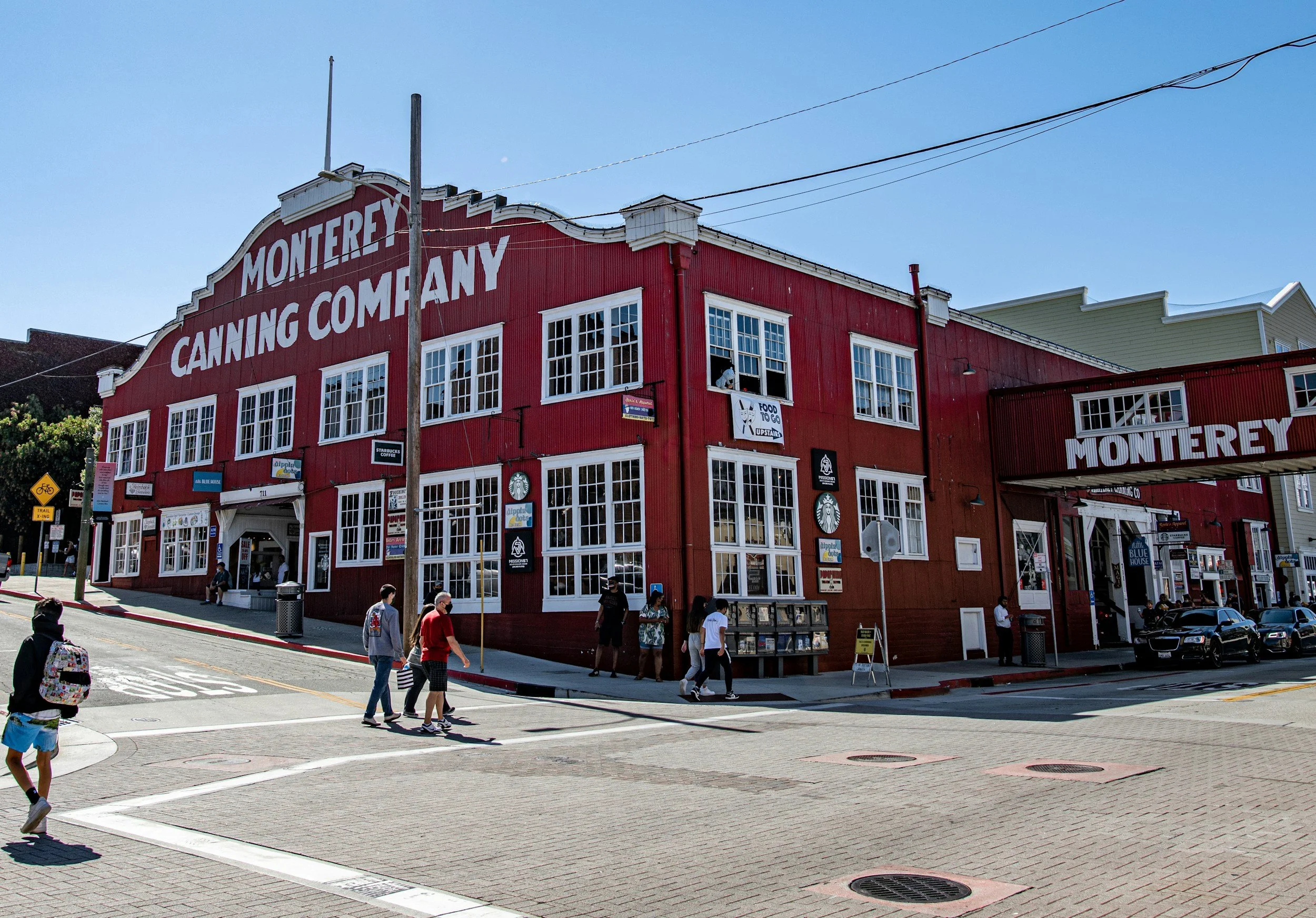 A red building with large white letters reading 'Monterey Canning Company' on the roof. Multiple windows, some with signs, and several people walking on the sidewalk in front. A crosswalk and parked cars are visible on the street in front.