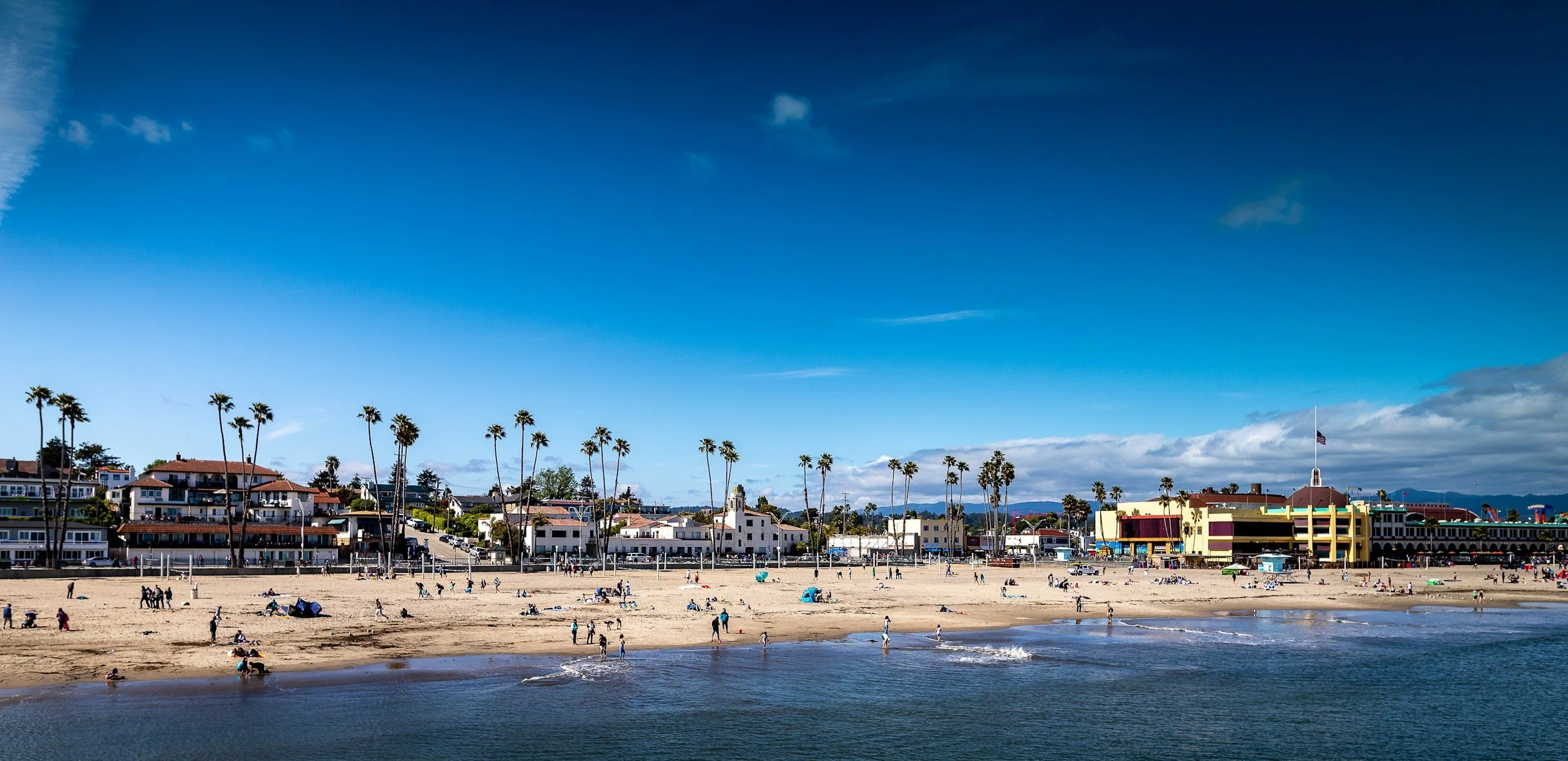 A distant view of a beach in Santa Cruz, California showing palm trees, water, and the sand populated with beach-goers.