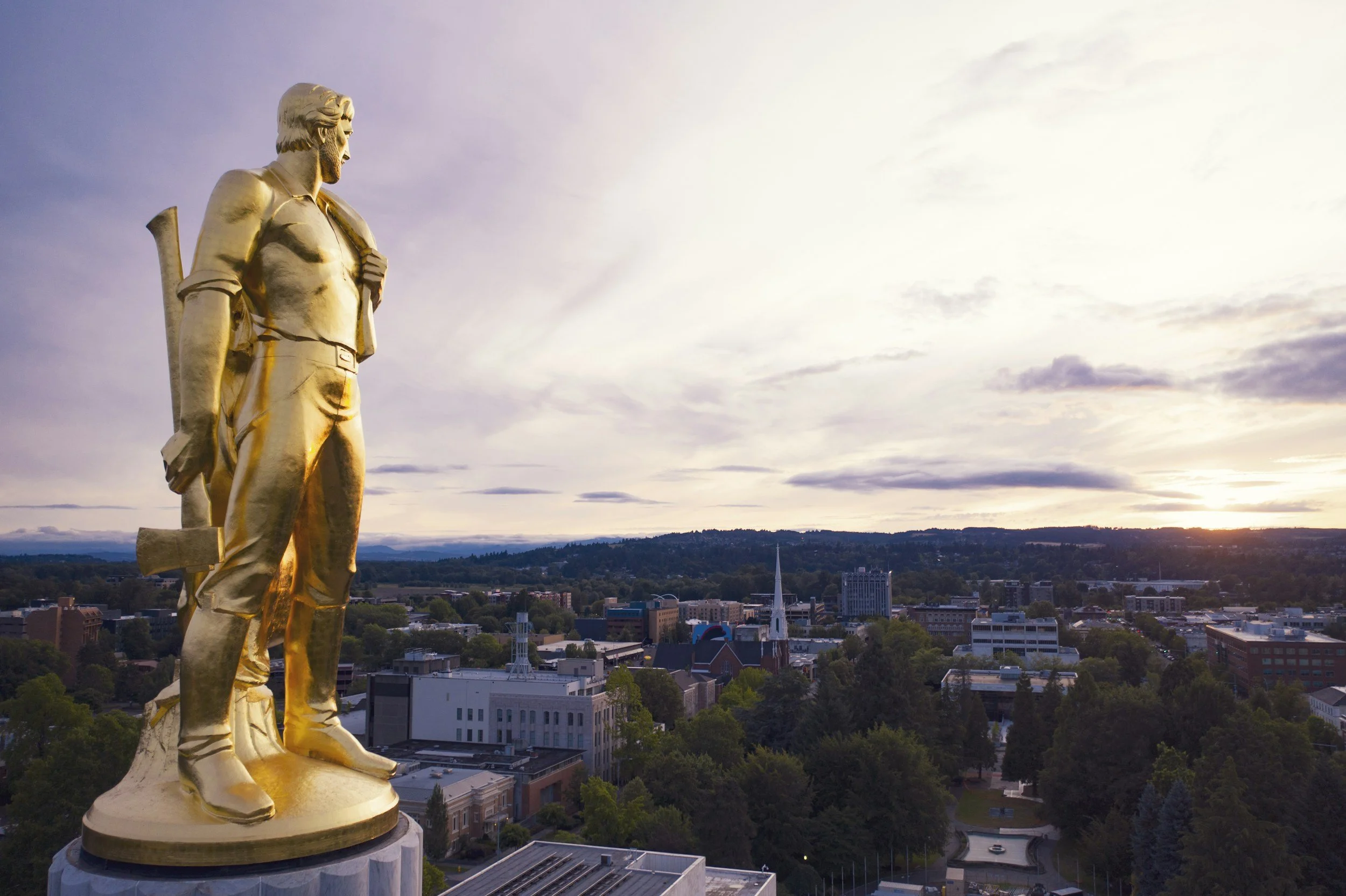 Gold statue of a man holding an axe, overlooking a cityscape at sunset on top of the Oregon State Capitol in Salem, Oregon.