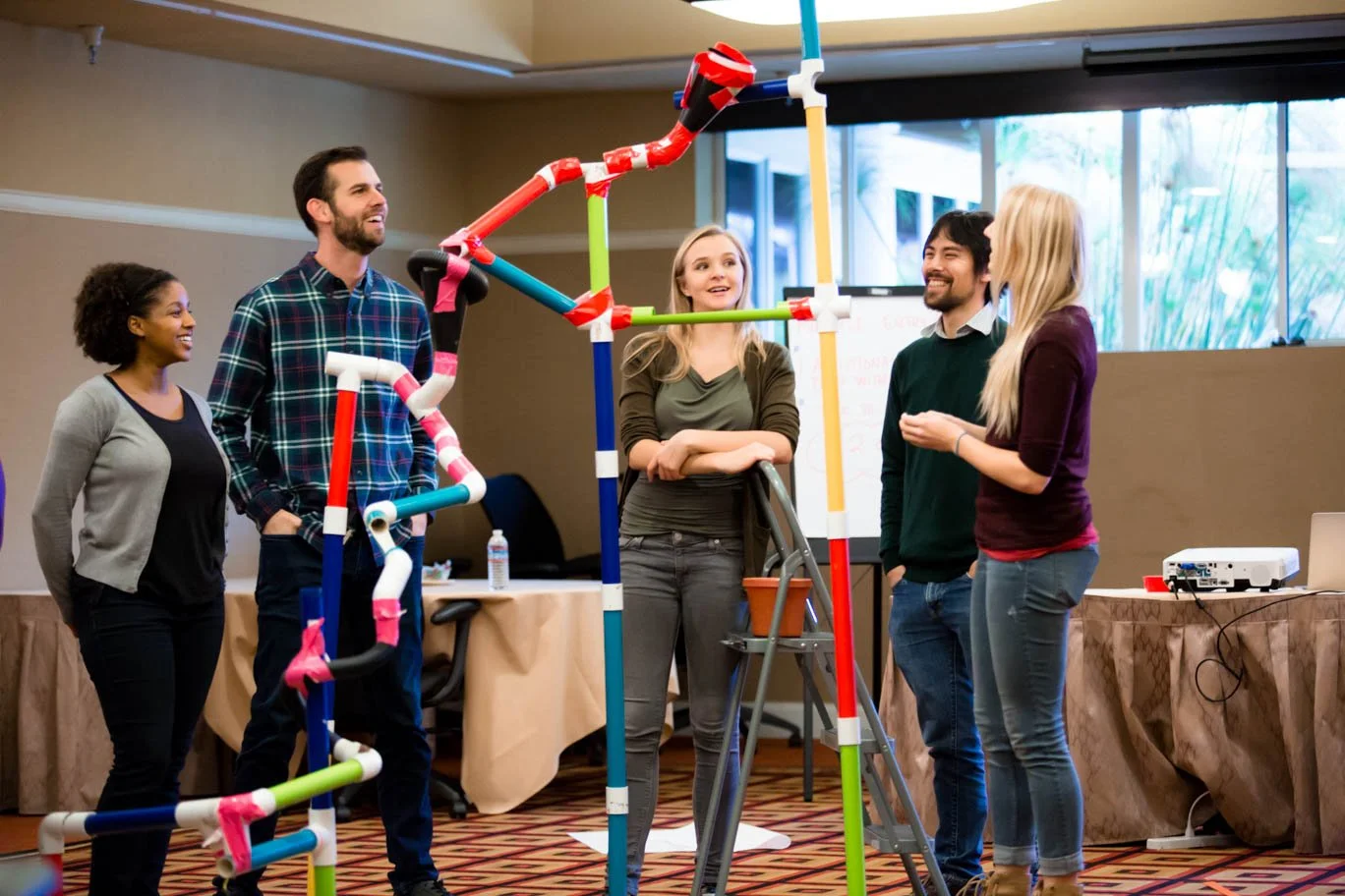 Group of five people in a meeting room, engaging in a team building activity involving a colorful pipe structure.