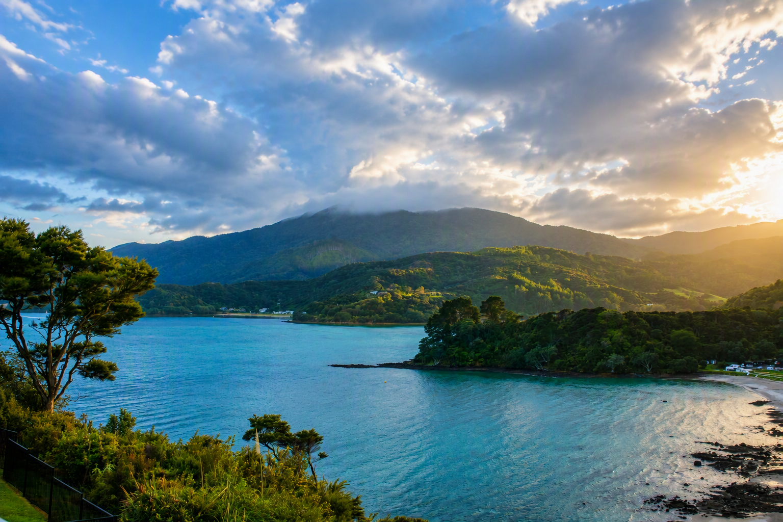 "Sunlight breaks through storm clouds over Long Bay, illuminating the lush hills of the Coromandel Peninsula as viewed from Coromandel Seaview Motel."