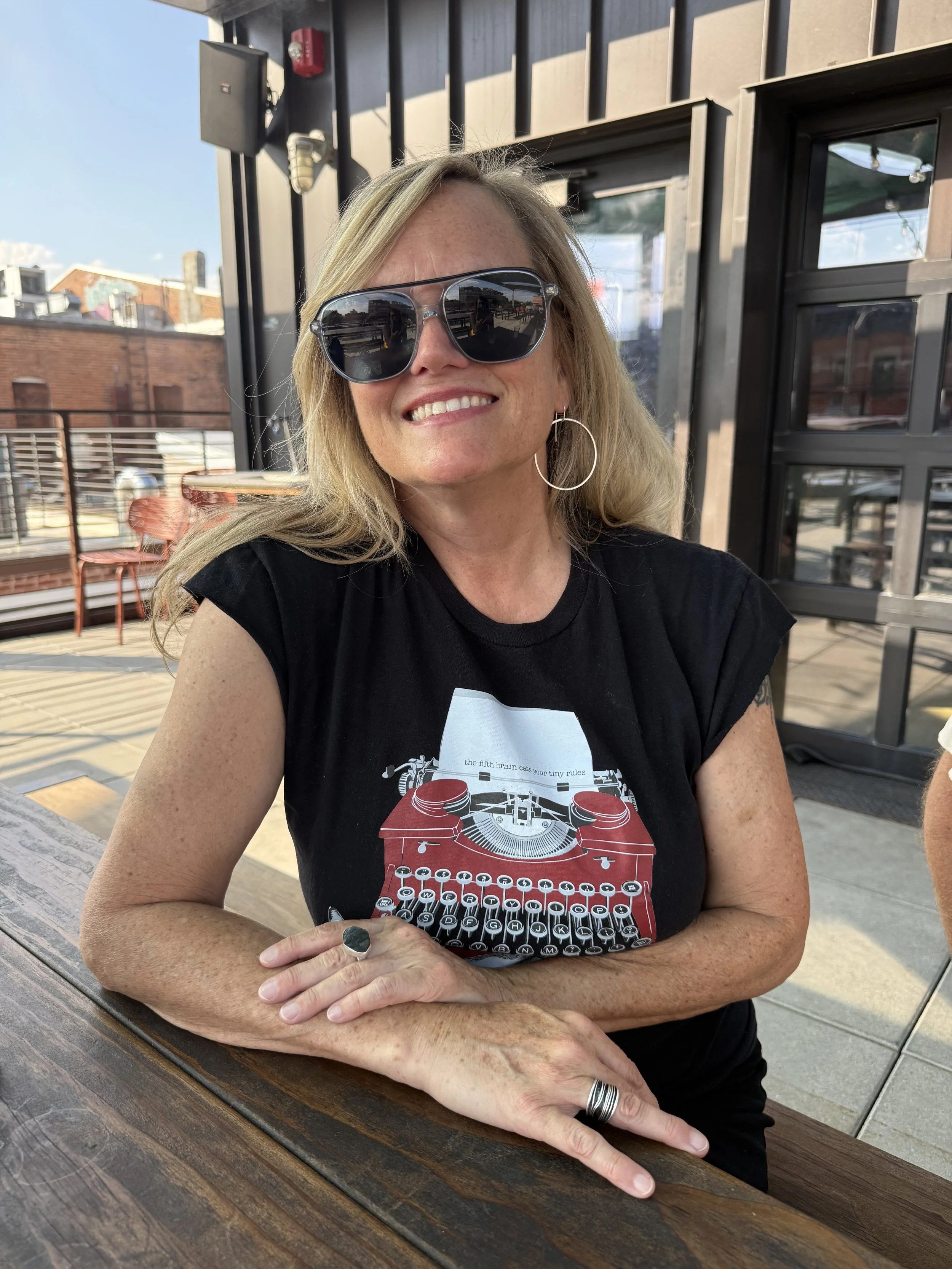 Frankie Rollins smiling in sunglasses and a black t-shirt with a red typewriter, sitting at a table in front of a building