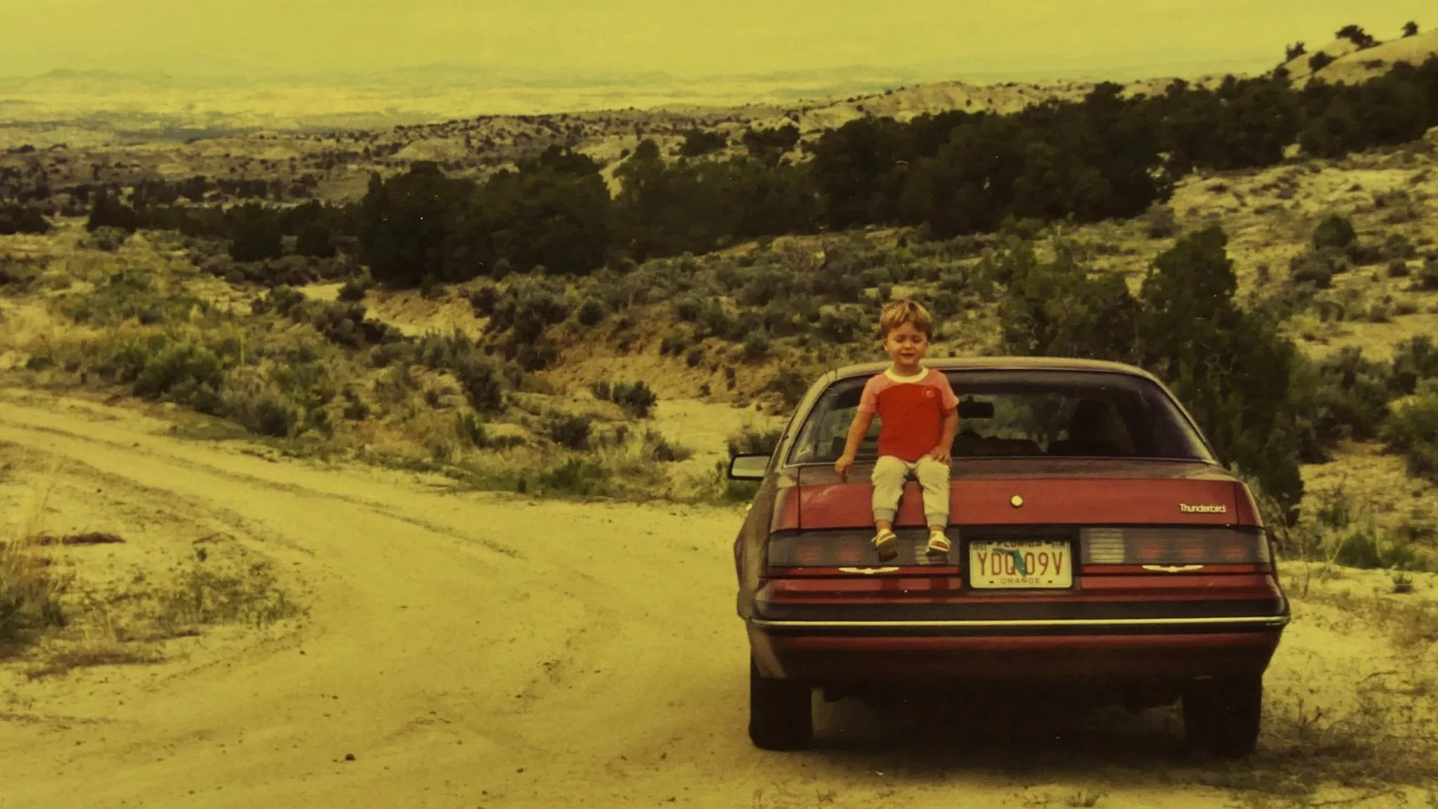 Criança sentada na traseira de um carro vermelho em uma estrada de terra em uma área de campo com vegetação e árvores ao fundo.