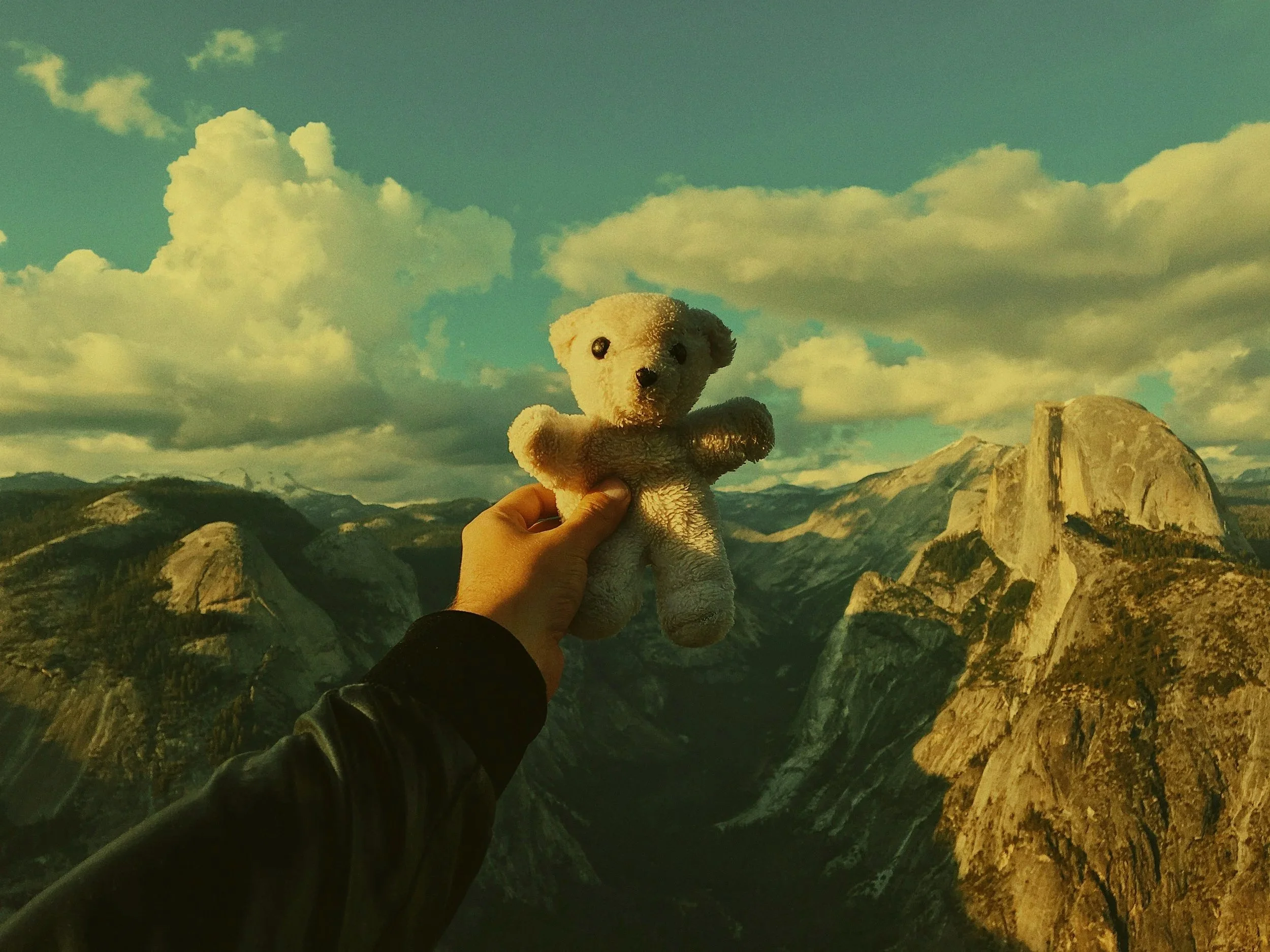 Mão segurando um ursinho de pelúcia branco na frente de uma paisagem de montanhas e céu com nuvens.
