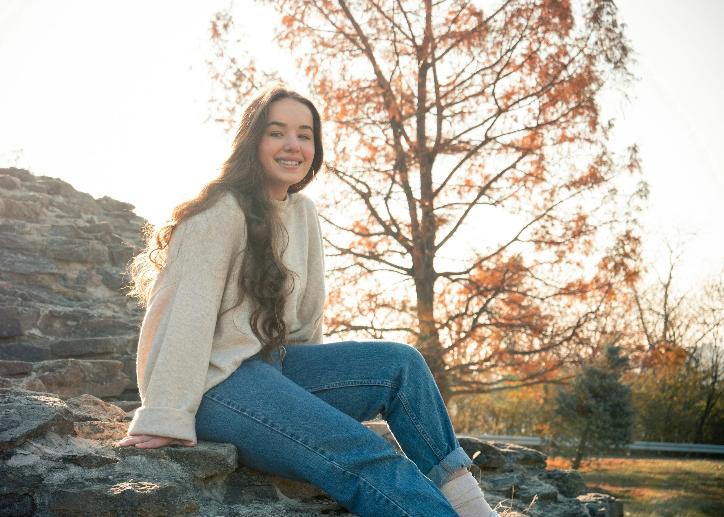 Smiling person sitting on rocks with autumn tree in background.