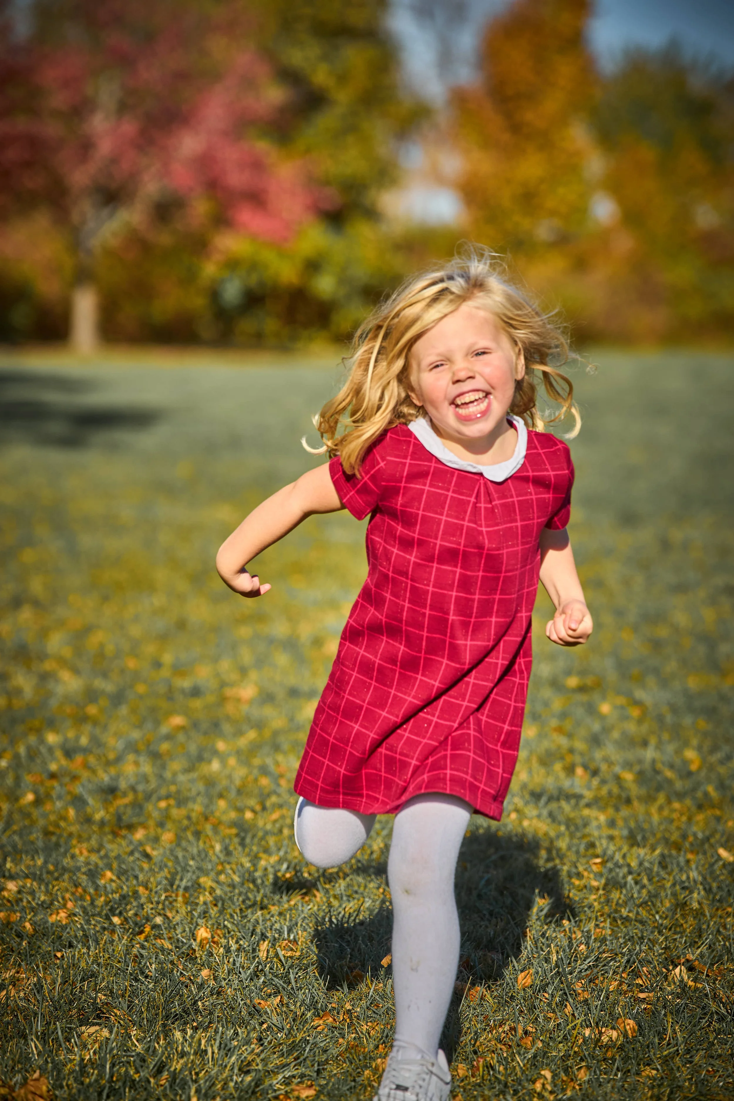 young girl in red dress runs through green field on autumn day big smile bouncing hair photograph by Life Times Photography