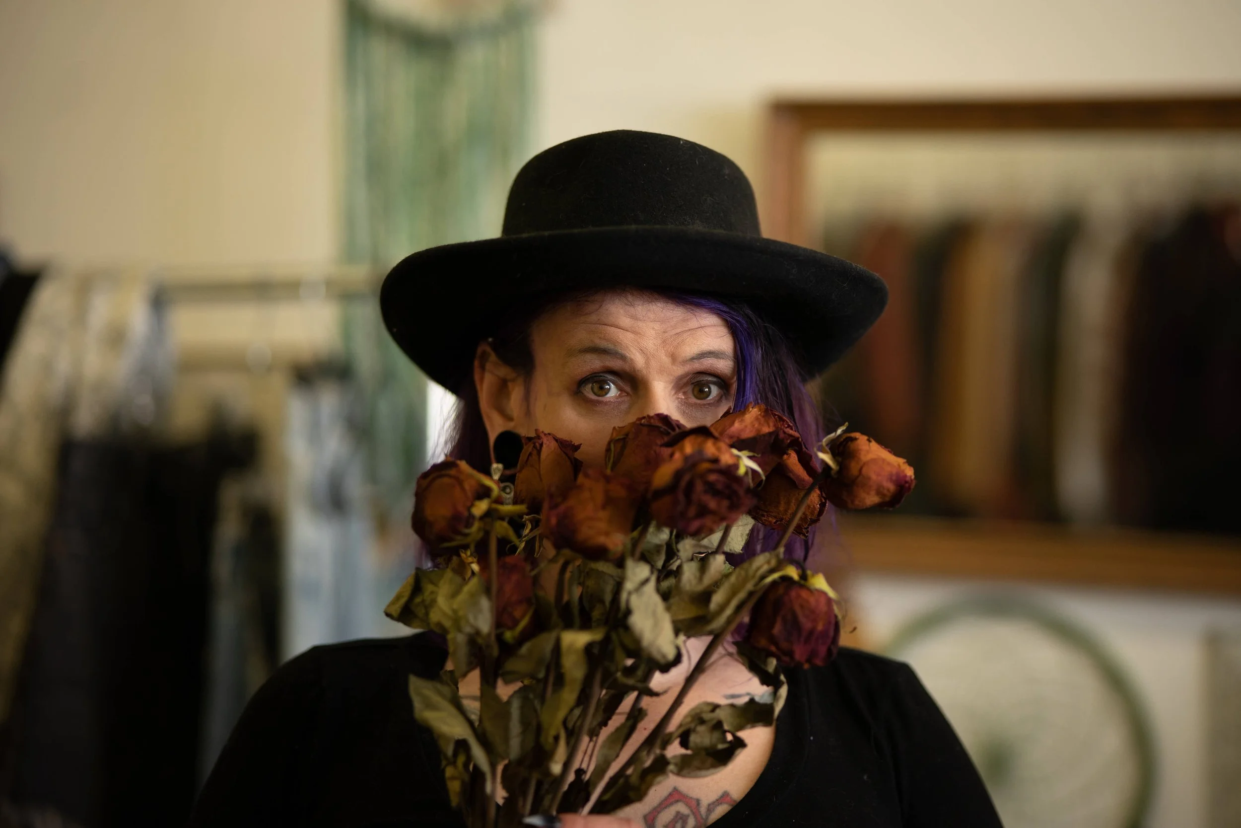 woman in black hat with purple hair peeking from behind bouquet of dried roses