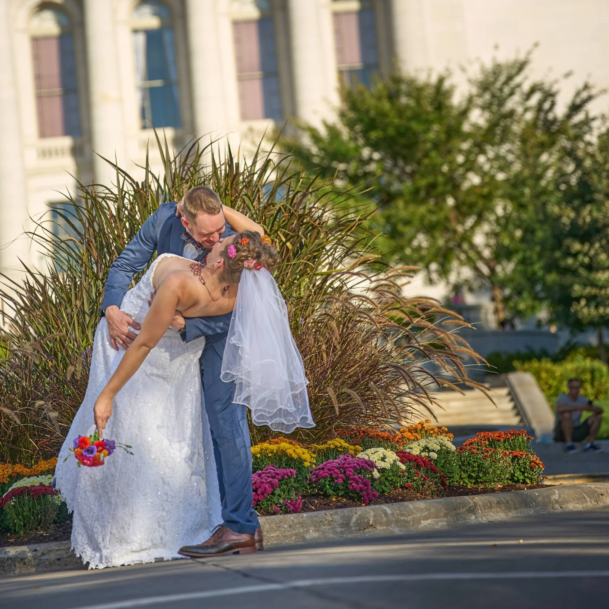 Bride and groom kissing outdoors near colorful flowers and greenery, with a building in the background.