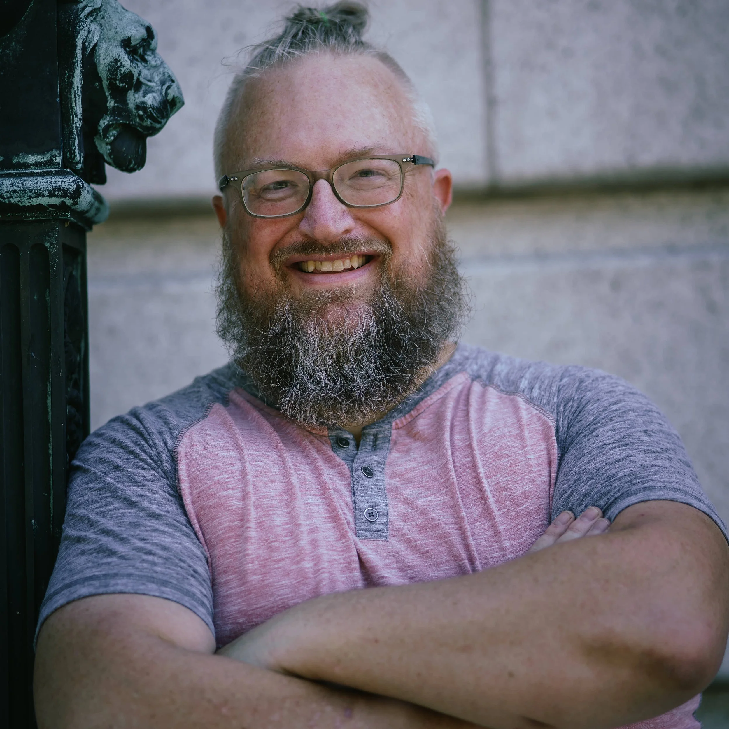 bearded man in pink and gray t-shirt poses with arms crossed, warm smile
