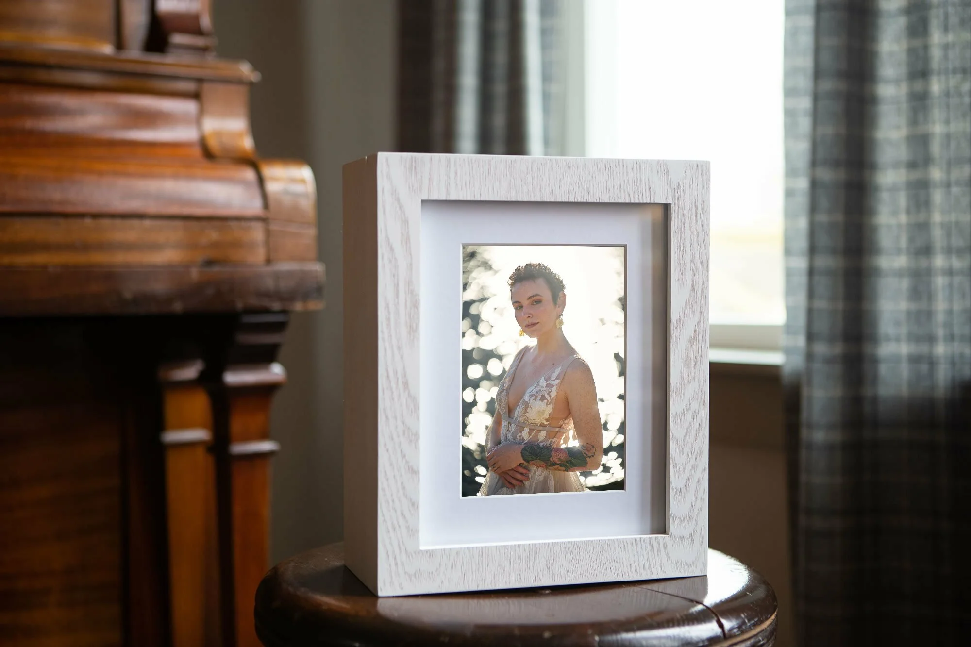 Framed photograph of a person with short hair and tattoos, placed on a wooden stool next to a piano in a room with gray curtains and a window.