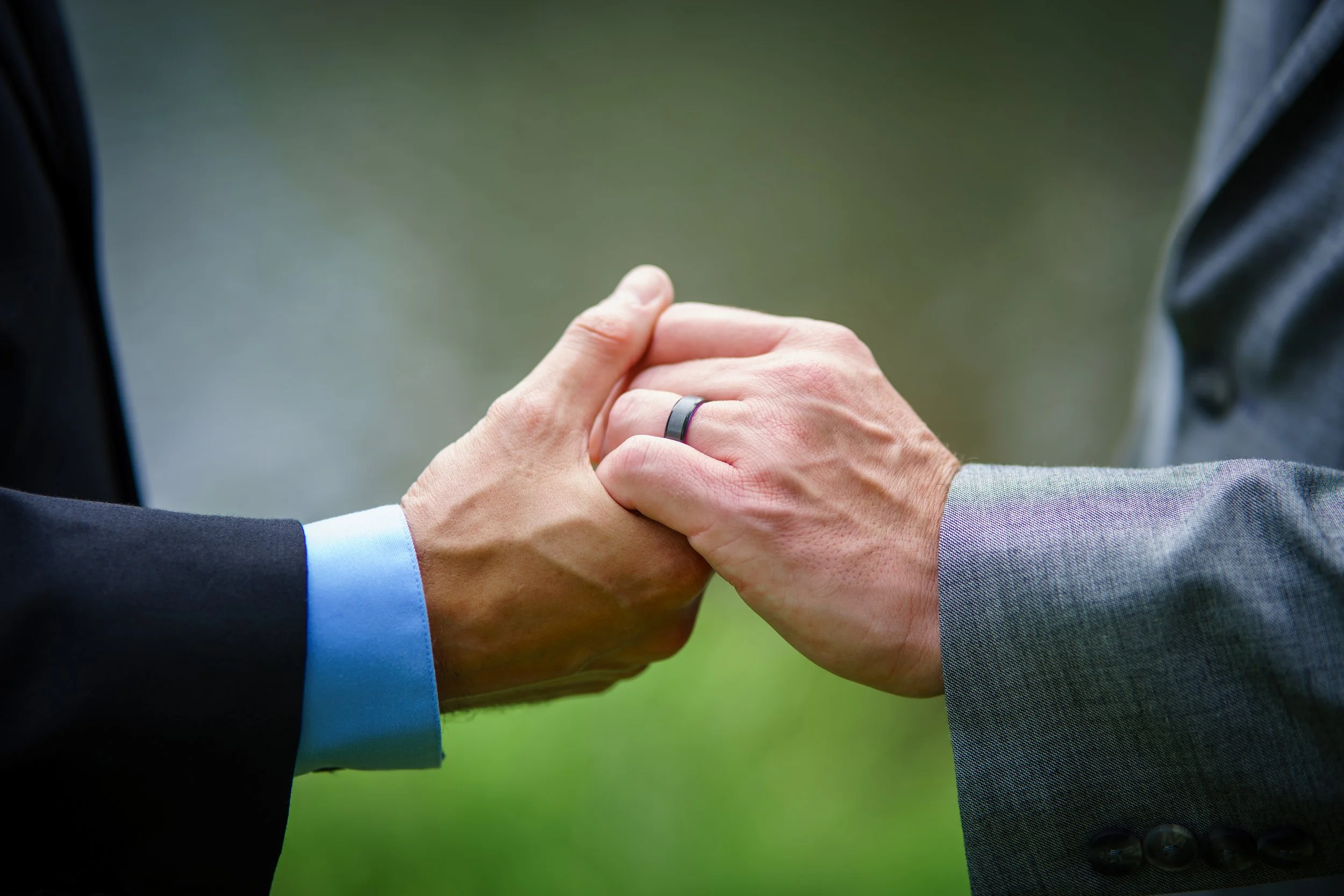 Close-up of two people in suits holding hands with a wedding ring visible
