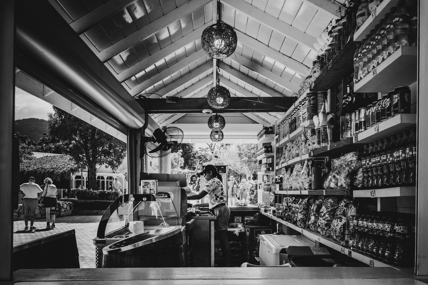 View inside a small store or café with a woman at the counter preparing an order. Shelves stocked with snacks and beverages on the right, and an outdoor scene with trees and people outside through the open window.