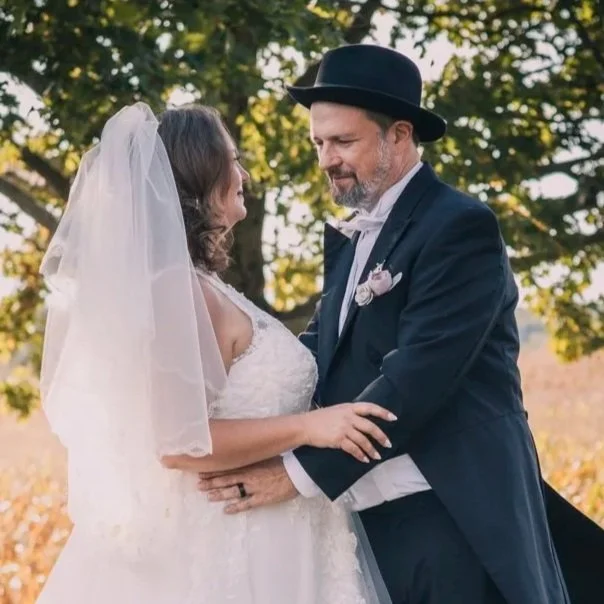 A bride and groom share a moment outdoors under a tree, with the groom wearing a black hat and suit, and the bride in a white wedding dress with a veil.