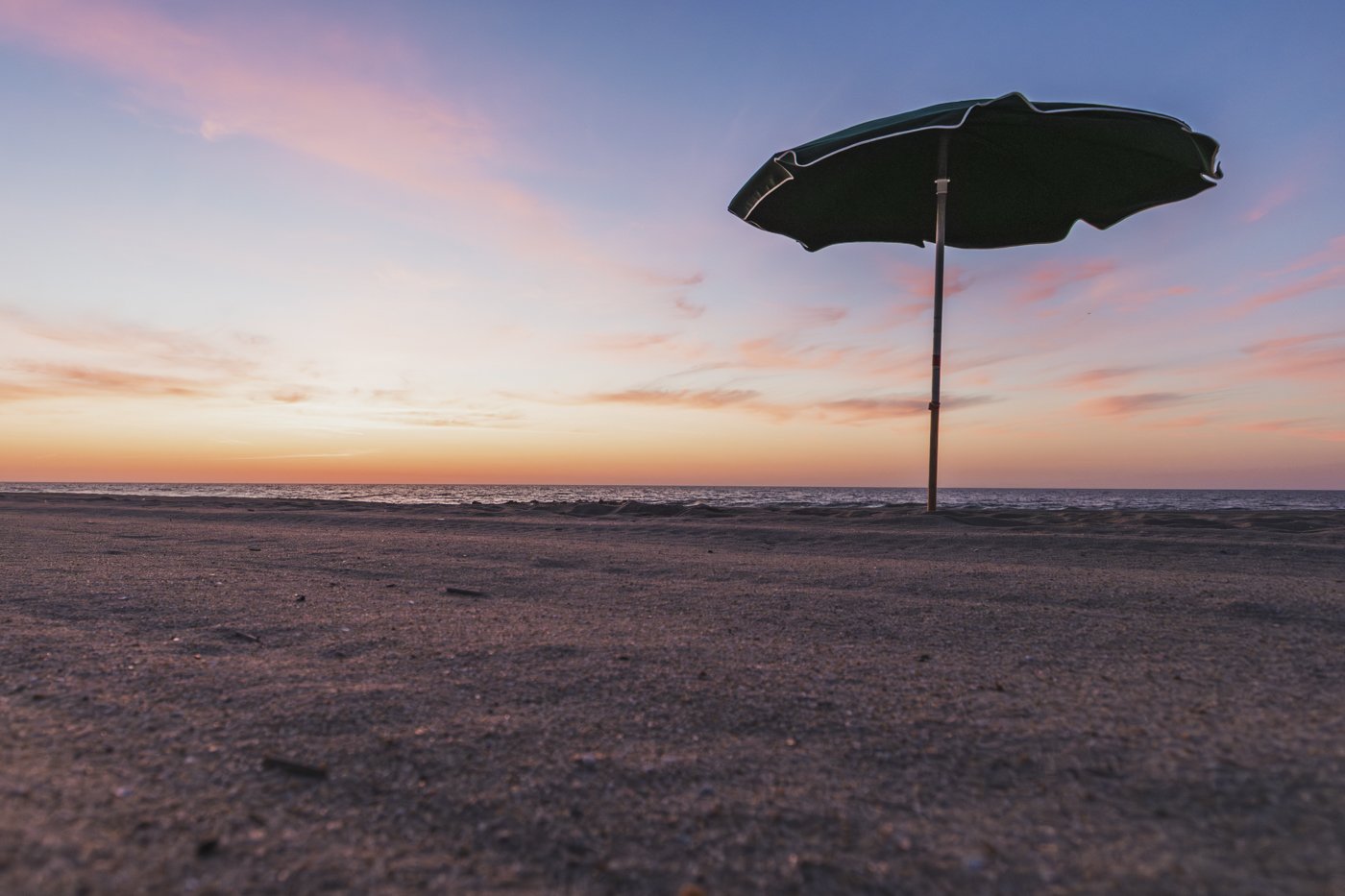 A beach scene at sunset with pink and orange sky, an empty black beach umbrella standing on the sand.