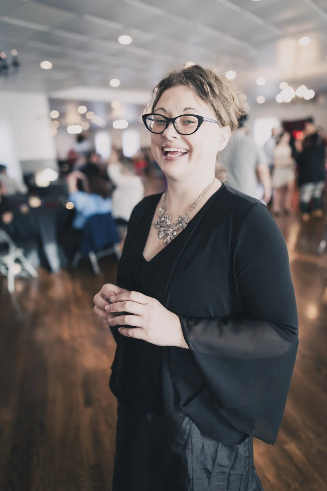 A woman with glasses and jewelry smiling in a dimly lit banquet hall filled with people and decorated with warm lights.