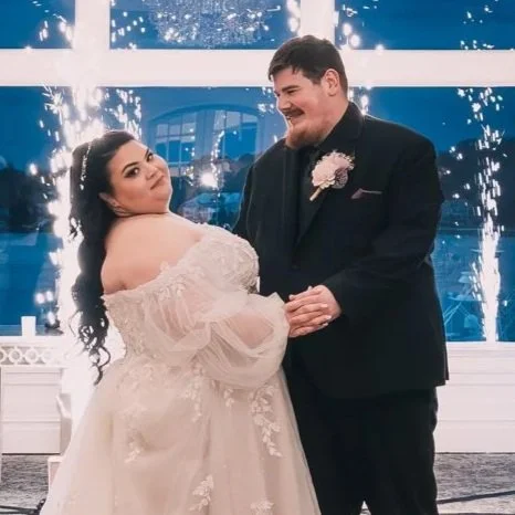 A bride and groom dance at their wedding reception, with sparklers in the background and big windows revealing a nighttime scene outside.