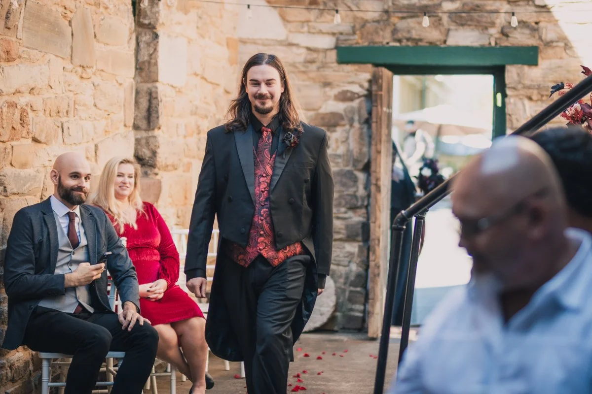 A man with long hair and a beard, dressed in a black suit with a patterned red vest, walks down an aisle at a wedding ceremony inside a rustic stone building. Seated behind him are a man in a suit holding a phone and a woman in a red dress, both watching him walk.