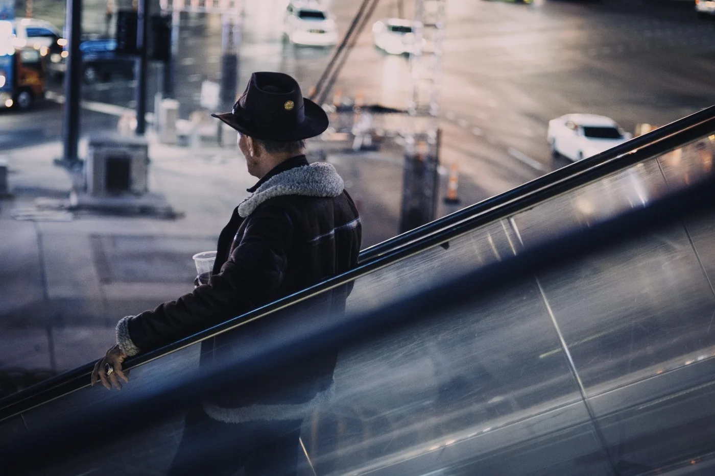 A man wearing a black hat and a brown jacket with a shearling collar, holding a plastic cup, standing on an escalator in an urban setting with cars and streetlights in the background.
