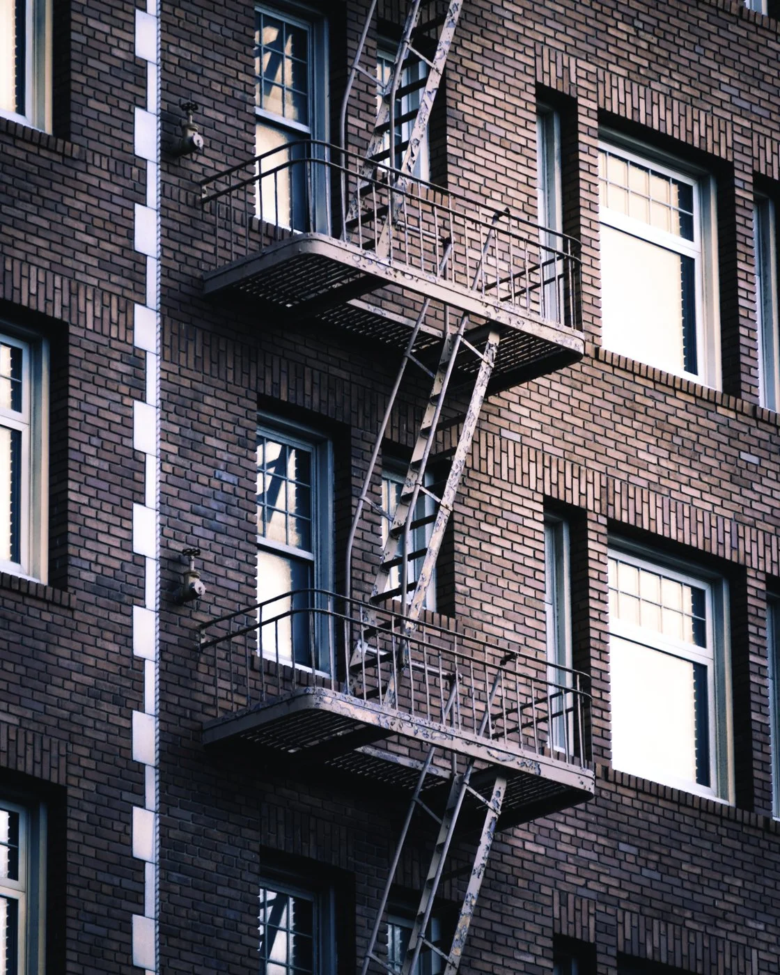 Close-up of a red brick apartment building with two fire escapes on the exterior, one above the other, with metal railings and ladders.