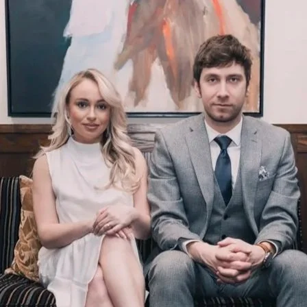 A woman in a white dress and a man in a gray suit sitting on a patterned sofa in front of abstract artwork.