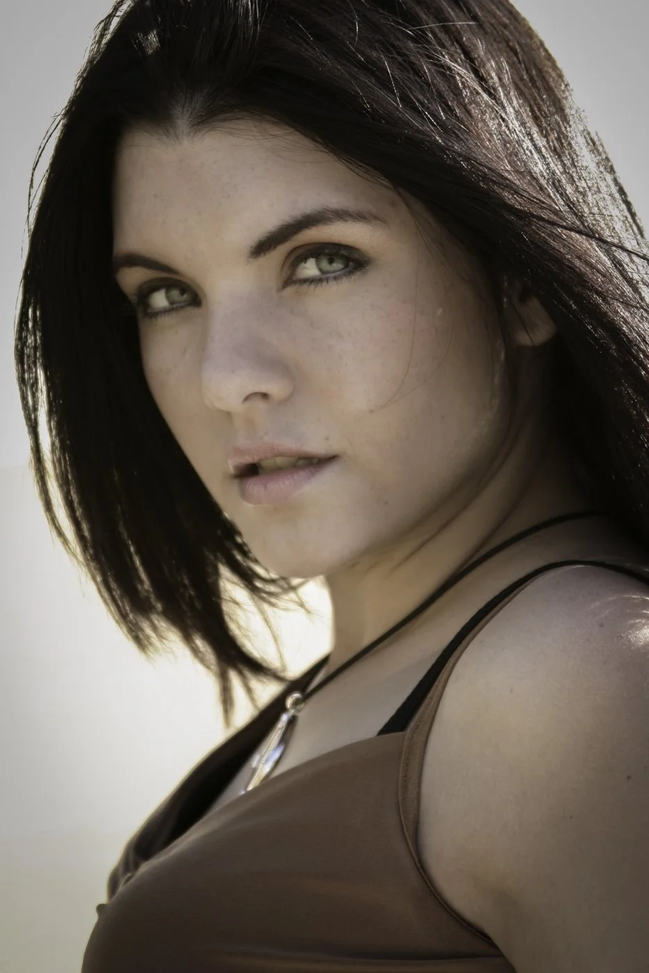 Close-up of a woman with dark brown hair, green eyes, and light makeup, wearing a brown top with black straps and a metal necklace.