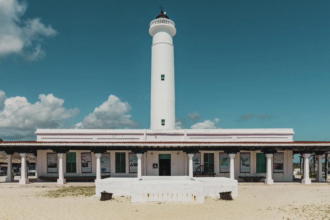 A tall white lighthouse with a red roof on top, situated behind a white building with a red-tiled roof, under a cloudy blue sky.