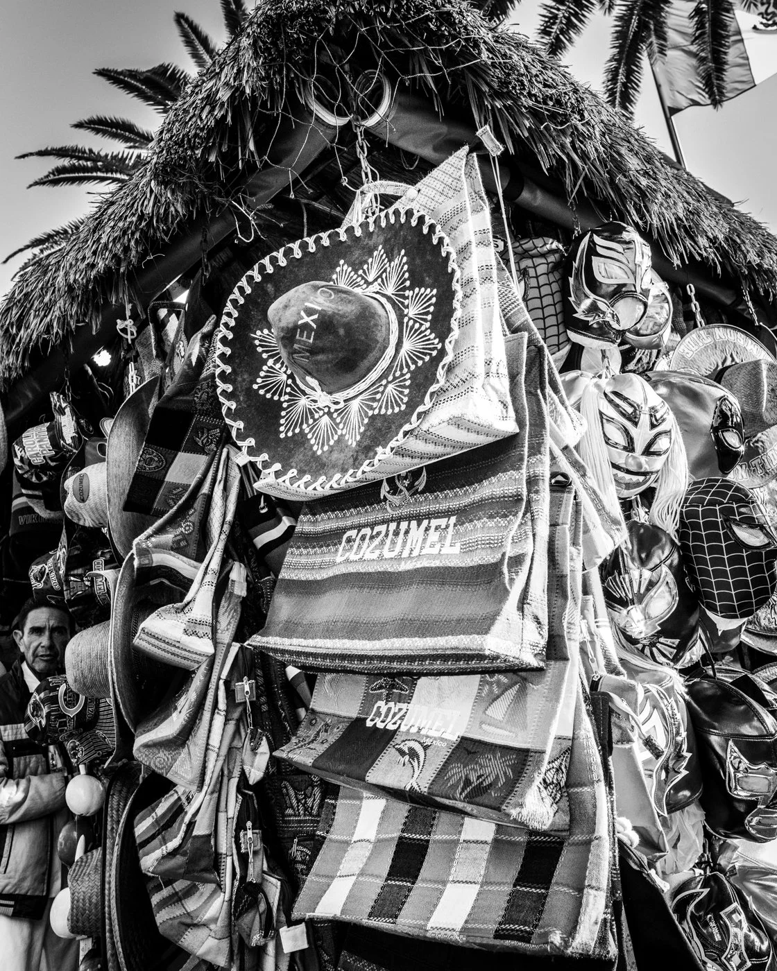 A display of various Mexican souvenirs including sombreros, masks, and woven textiles, with a palm tree and a flag in the background.