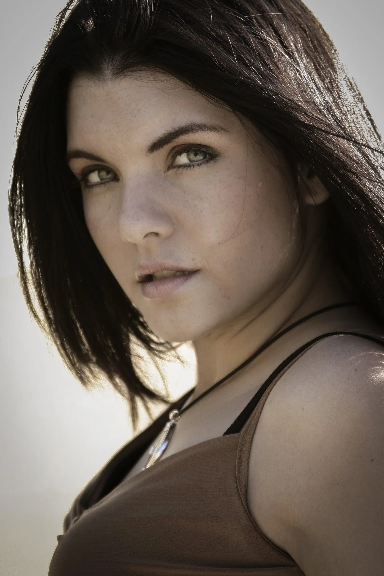 Close-up of a woman with dark hair, fair skin, and green eyes looking at the camera, wearing a brown top with thin straps and a black necklace.