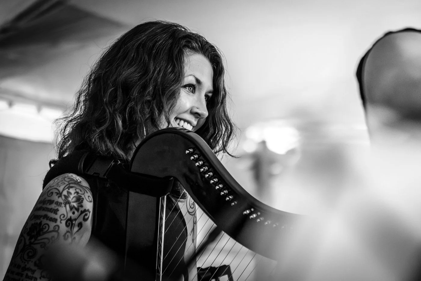 A woman with wavy hair, smiling, holding a harp, with tattoos on her arm, in a black-and-white photo.