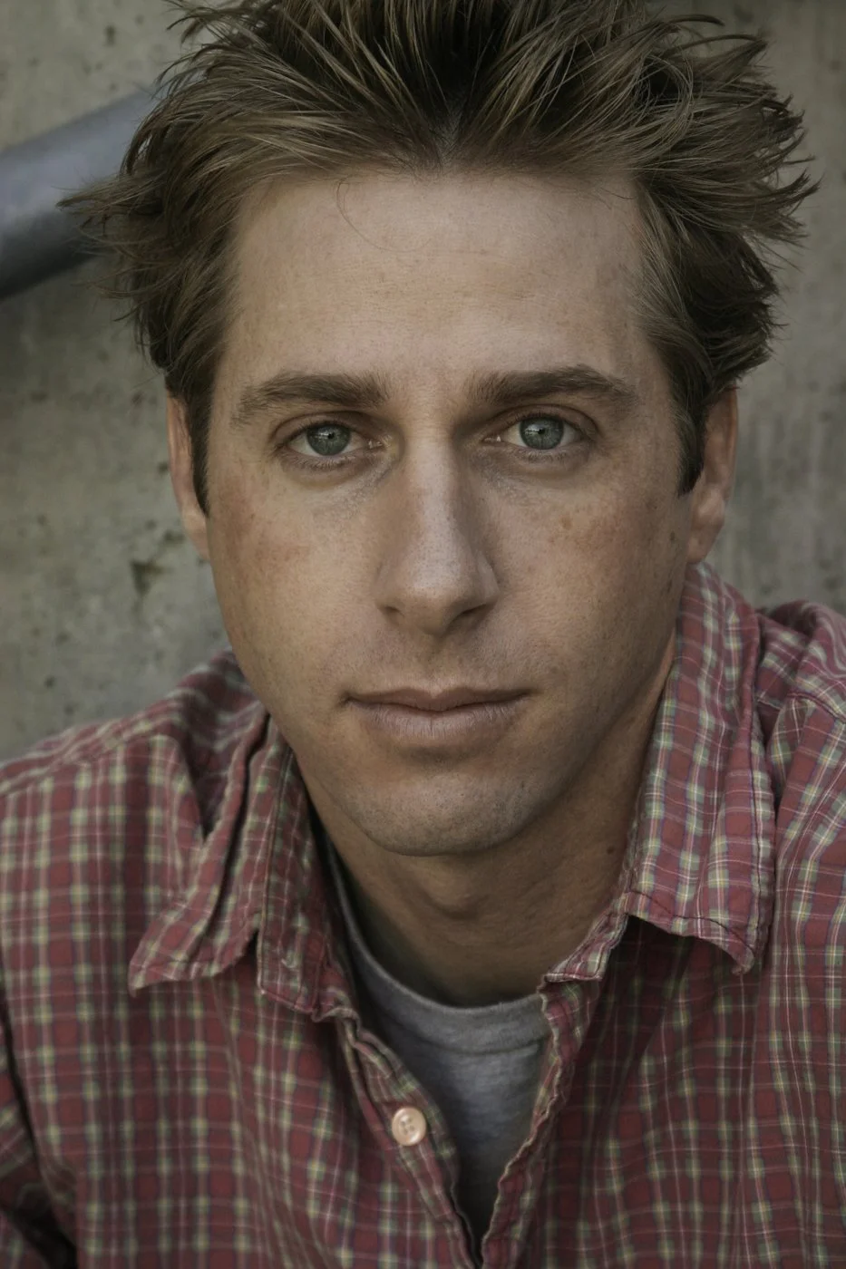 Close-up of a young man with blue eyes, light skin, and styled brown hair, wearing a red checkered shirt over a gray T-shirt, sitting outdoors with a concrete wall in the background.