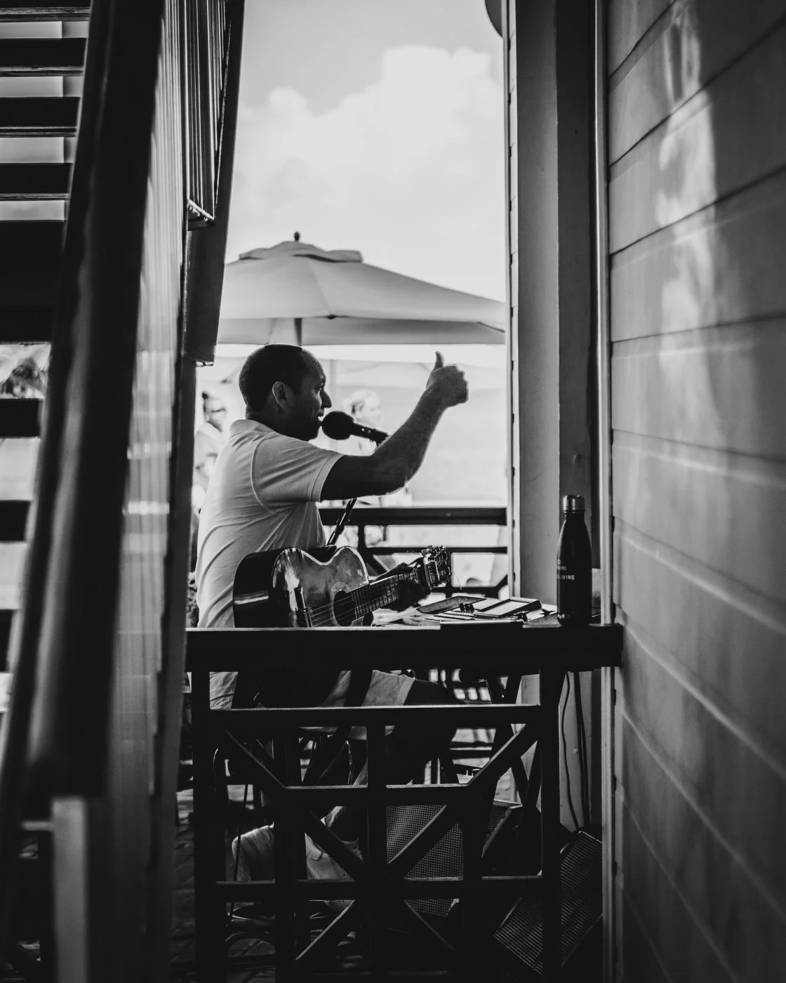 A man playing guitar and singing into a microphone at an outdoor setting. He is giving a thumbs-up gesture. There are people in the background under an umbrella, with a wooden wall on the right side.